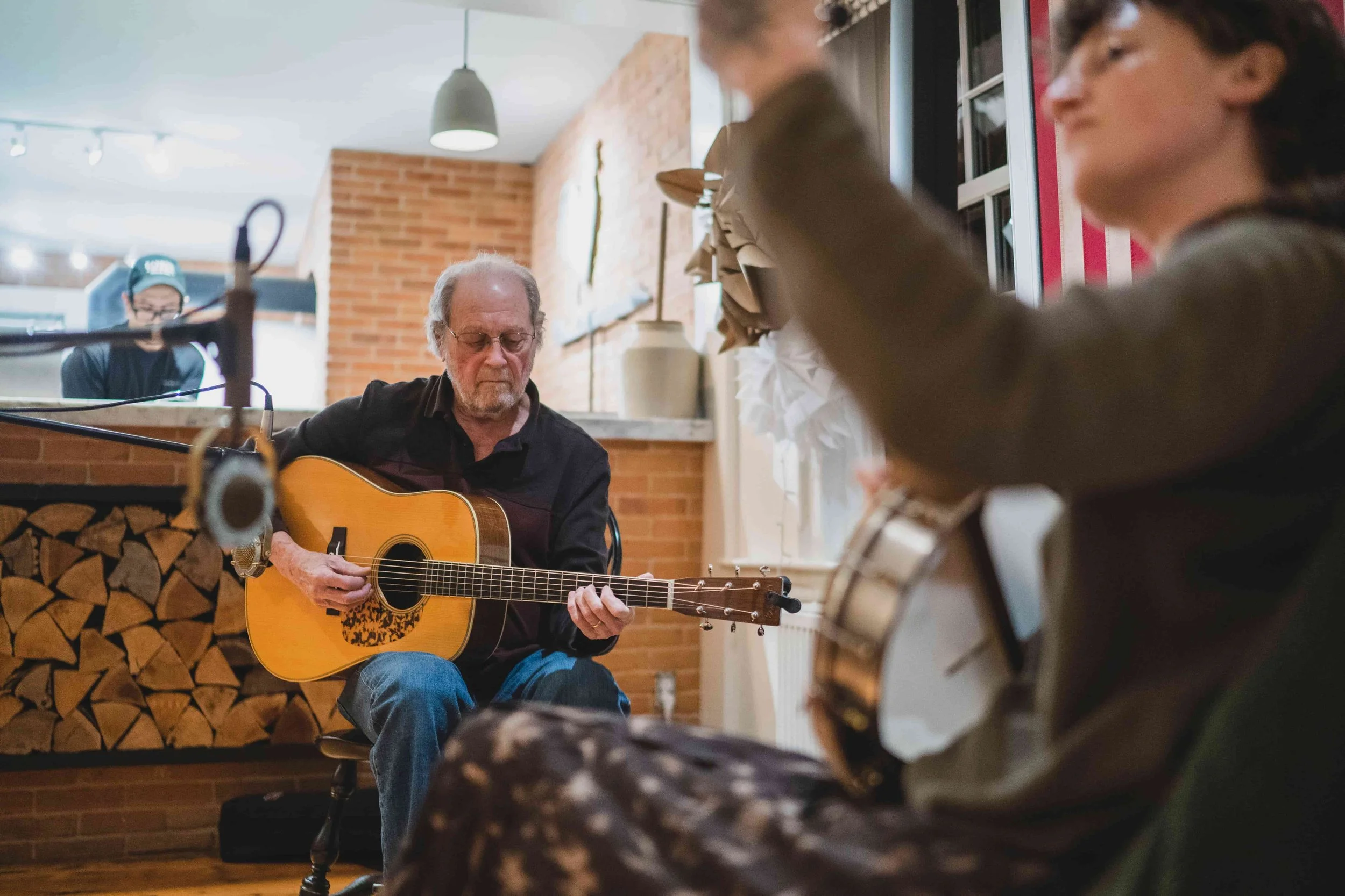 An elderly man playing an acoustic guitar while sitting on a stool inside a cozy, brick-walled room. There is a woman in the foreground with some papers, and a kitchen area with a person in a cap in the background.
