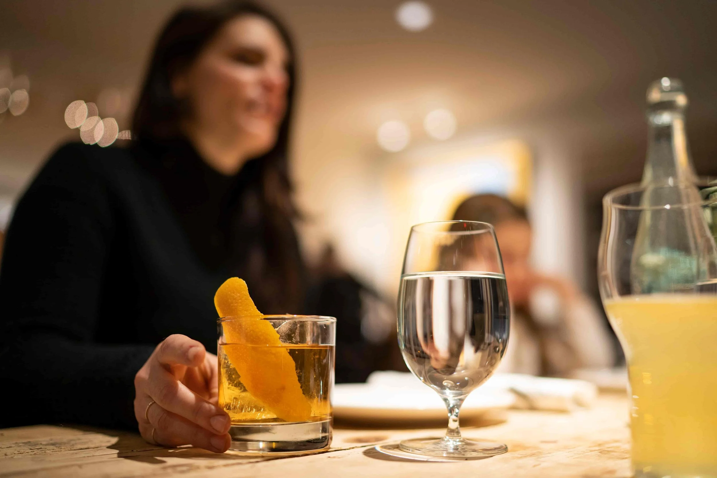 A woman holding a glass of water with a lemon twist in a restaurant setting, with a blurred background of lights and other glasses.