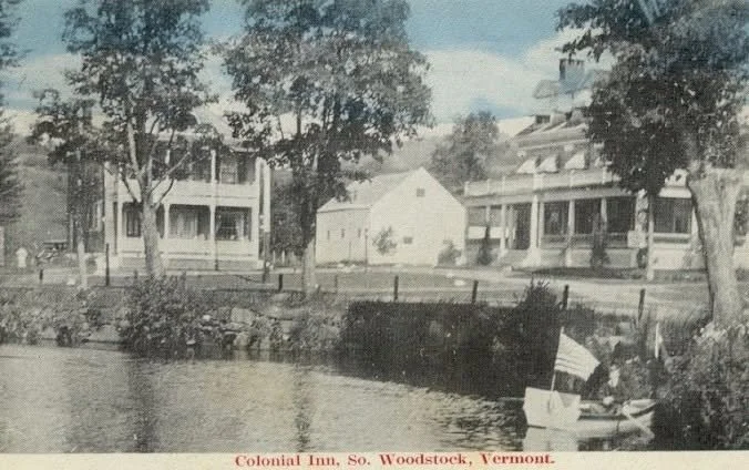 A black and white photo of a waterfront scene at the Colonial Inn in South Woodstock, Vermont, featuring buildings, trees, and a small boat near the shore.