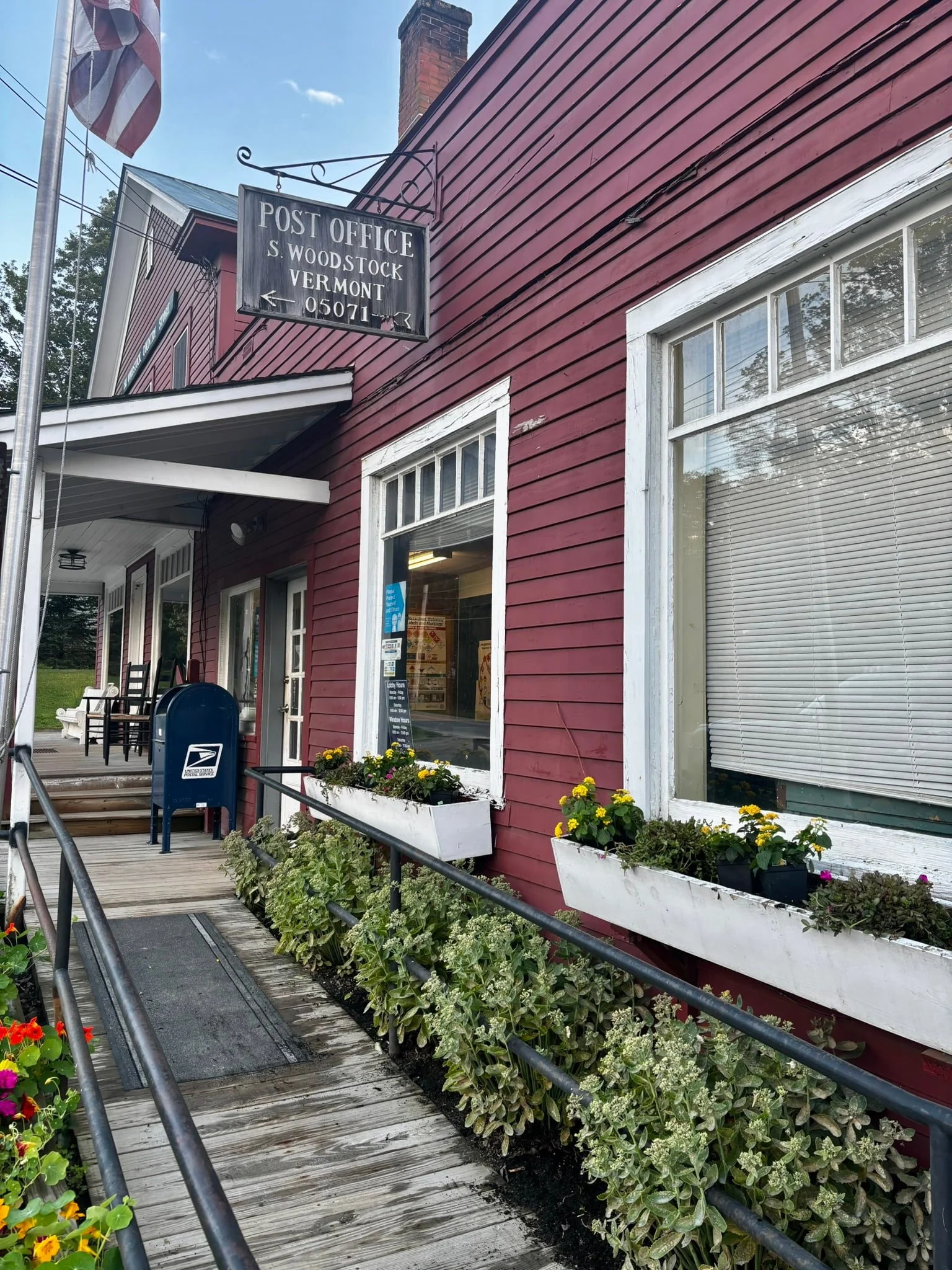 Red wooden building with white window trim and flowers in planters outside, USPS mailbox, chairs, flagpole with American flag, and a sign indicating a post office in Woodstock, Vermont.