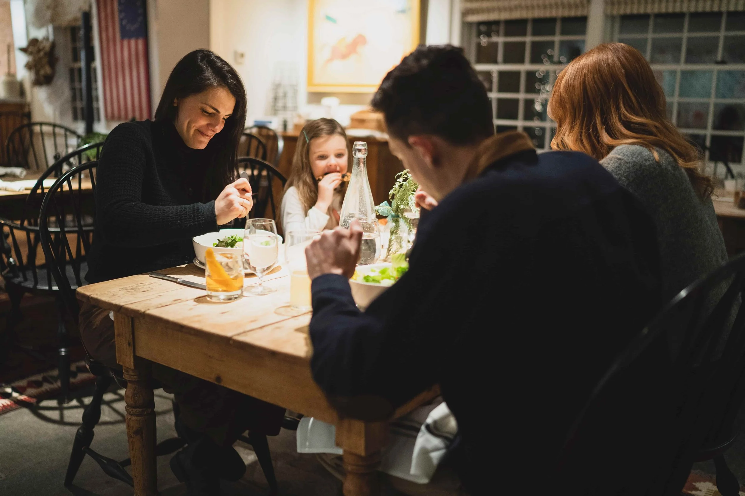 A woman and two children sitting at a dining table, enjoying a meal together in a cozy, warmly lit kitchen or dining room.