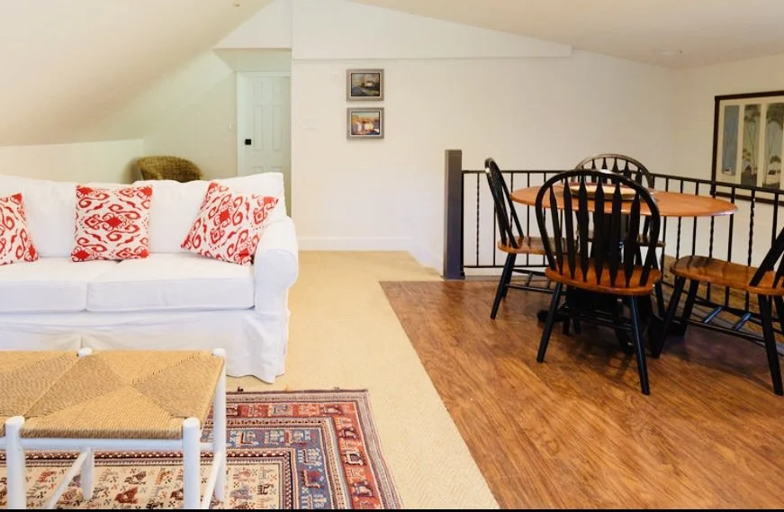 Living room with white sofa, red and white patterned cushions, wooden dining table with black chairs, hardwood and carpeted flooring, with paintings on the wall and a window.