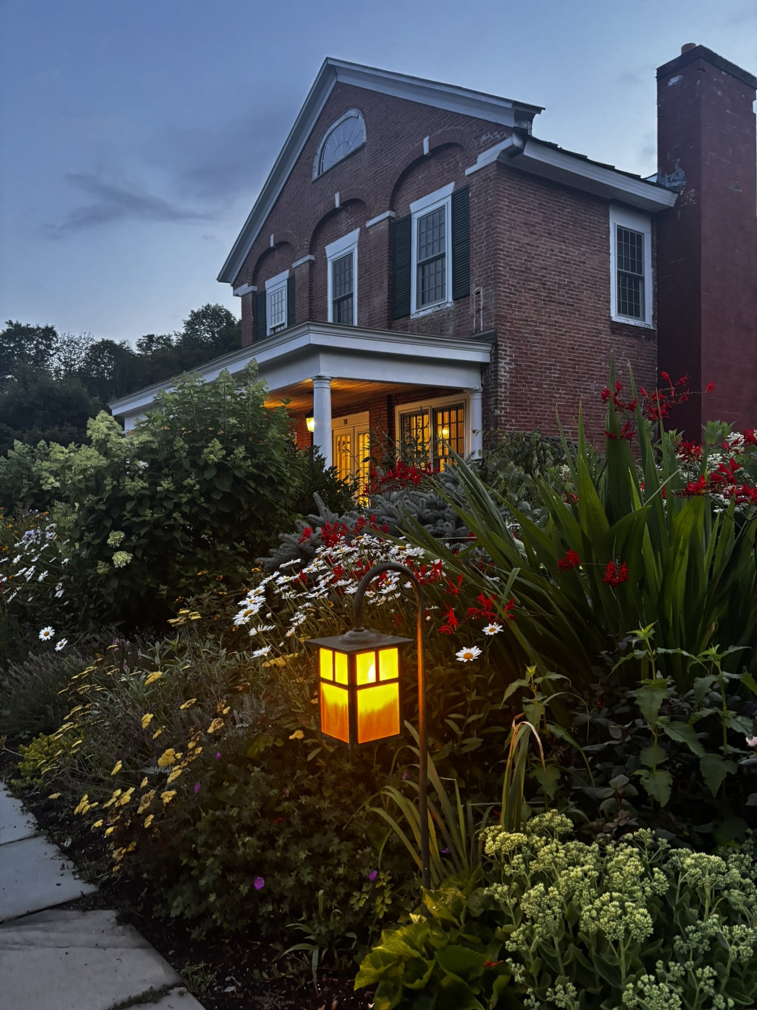 A large brick house with white-framed windows and a small porch with a light on, surrounded by a garden with various flowers and plants, illuminated by a glowing outdoor lantern at dusk.