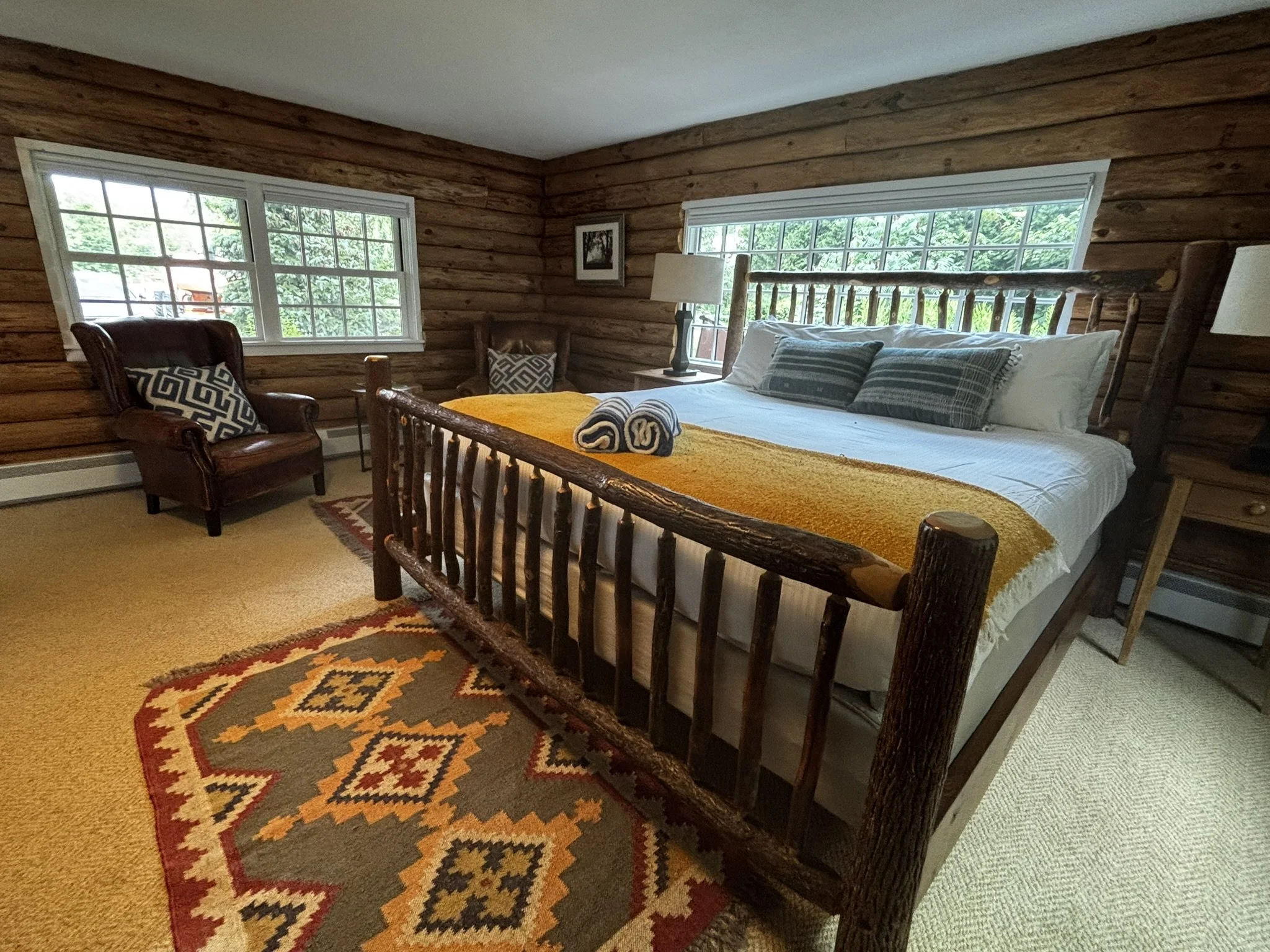 A cozy bedroom with log cabin style wooden walls, a wooden bed frame with white bedding and striped pillows, a leather armchair with decorative pillows, a bedside table with a lamp, large windows showing green trees outside, and a colorful patterned rug on the carpeted floor.