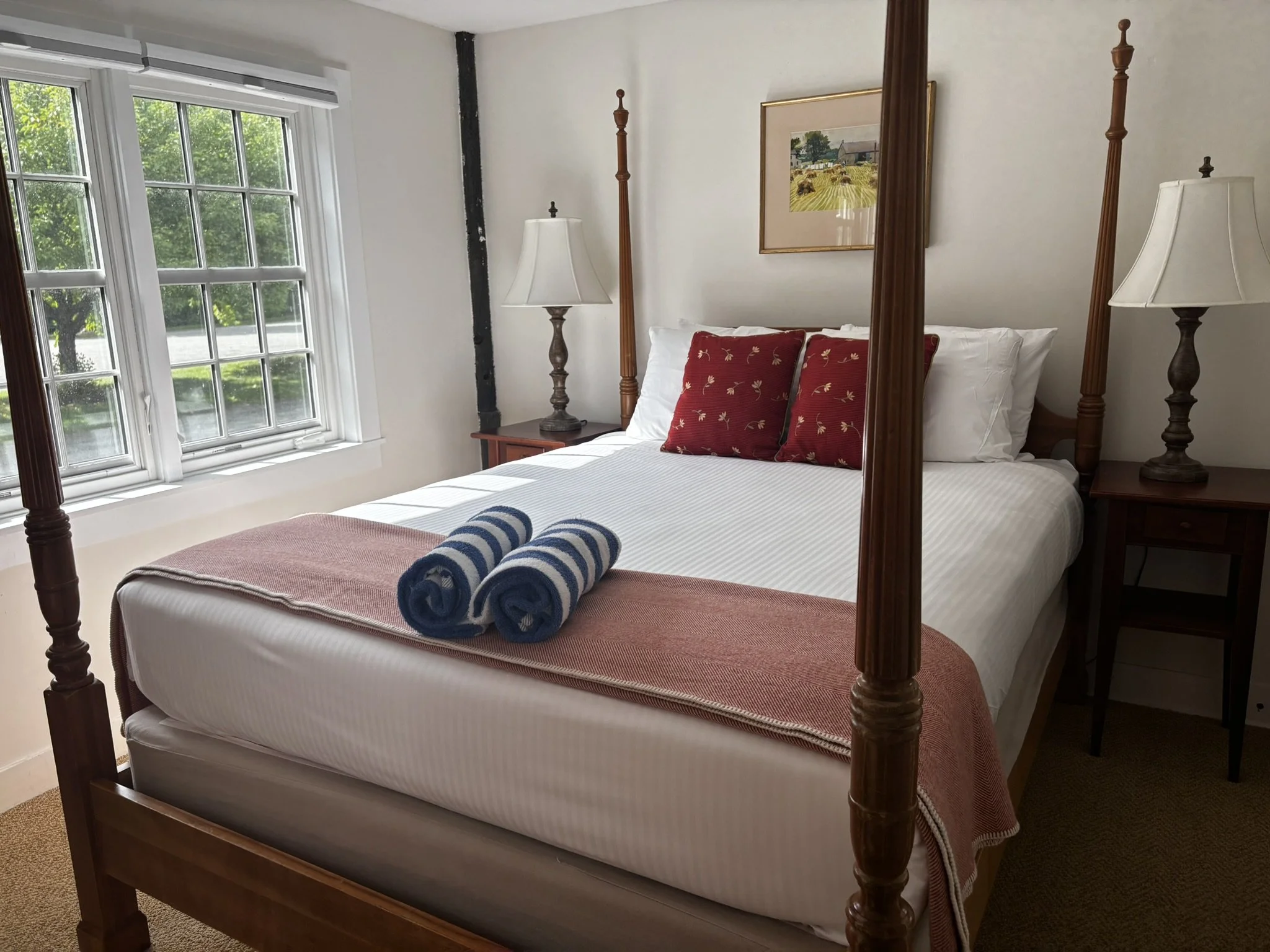 A bedroom with a four-poster bed dressed with white sheets, red decorative pillows, and a folded towel on the foot of the bed. There are two bedside tables with lamps and a window with multiple panes showing green trees outside.