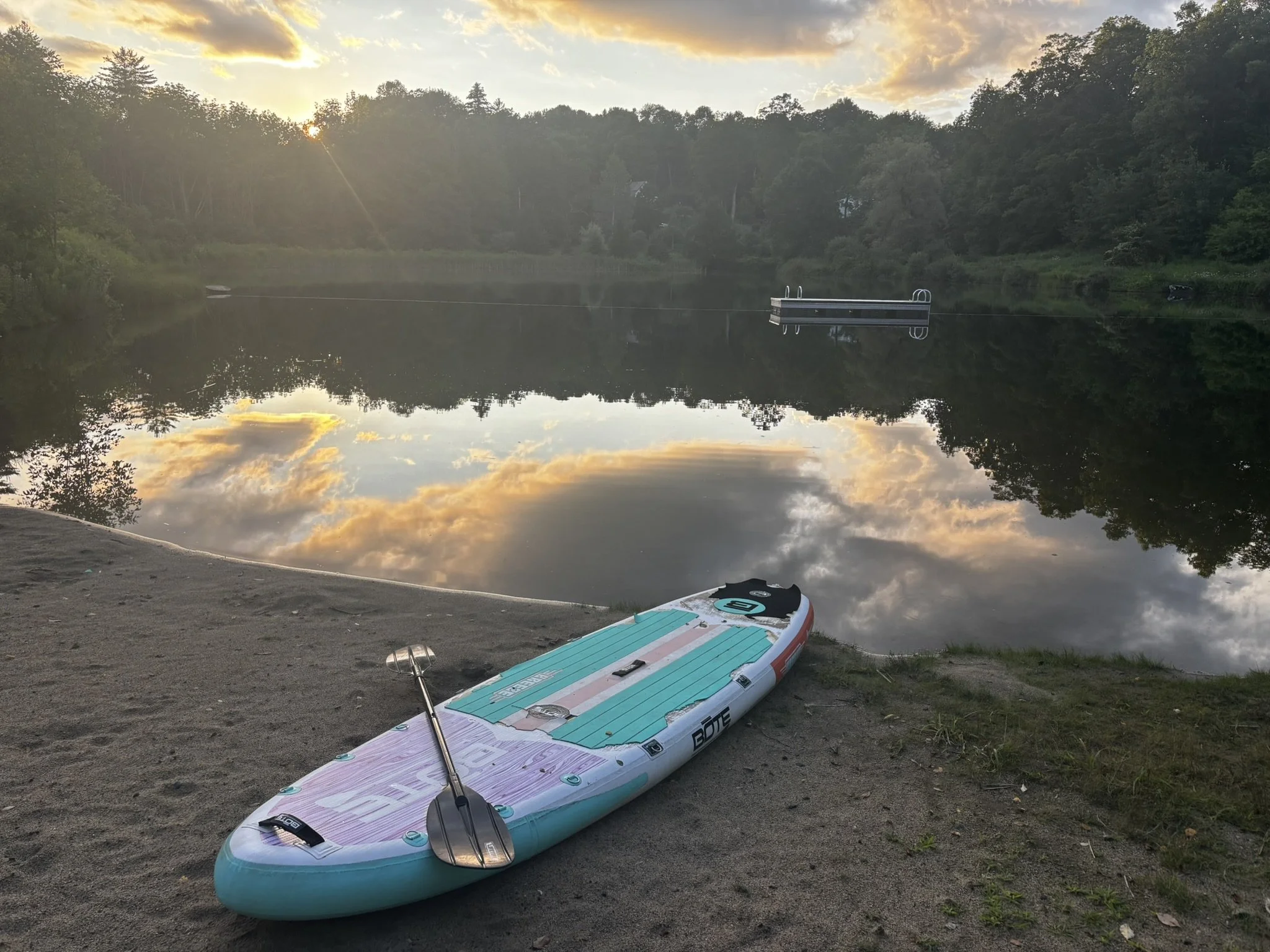 A paddleboard with a paddle resting on it, placed on a dirt shore by a calm lake during sunset or sunrise, with trees and sky reflected in the water.