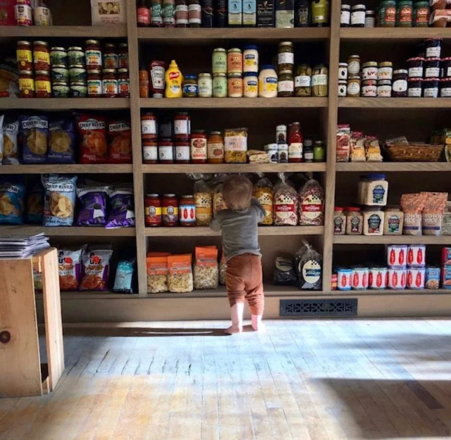 Young child standing on tiptoes and reaching up in front of grocery store shelves filled with canned and packaged food items.