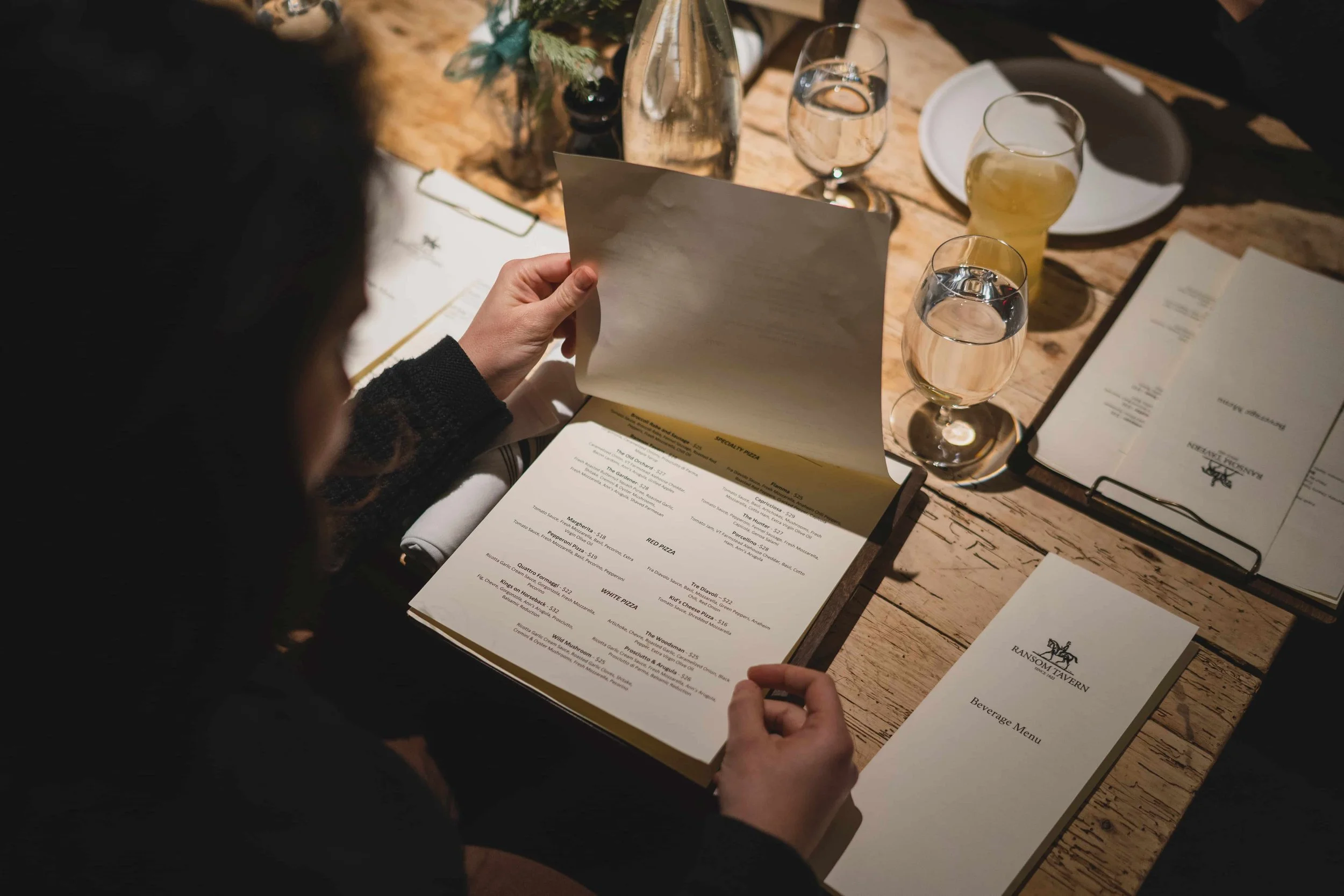 A person holding a restaurant menu at a wooden table with glasses of water, beer, and a water bottle, in a dimly lit dining setting.