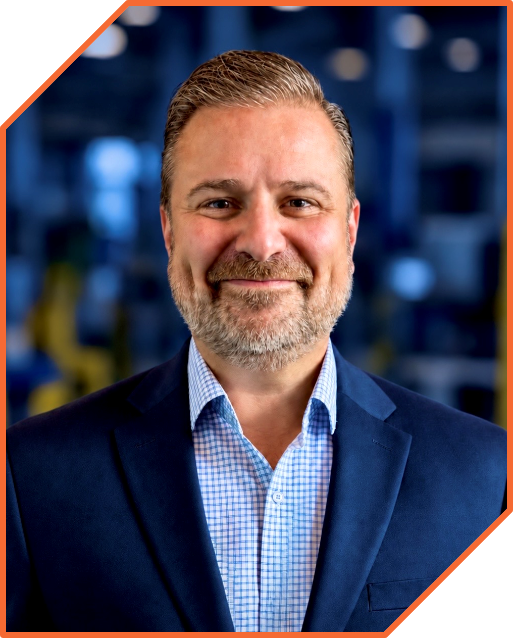 Portrait of a middle-aged man with a beard, smiling, wearing a blue suit jacket and a blue checkered shirt, with a blurred background of an industrial or tech environment.