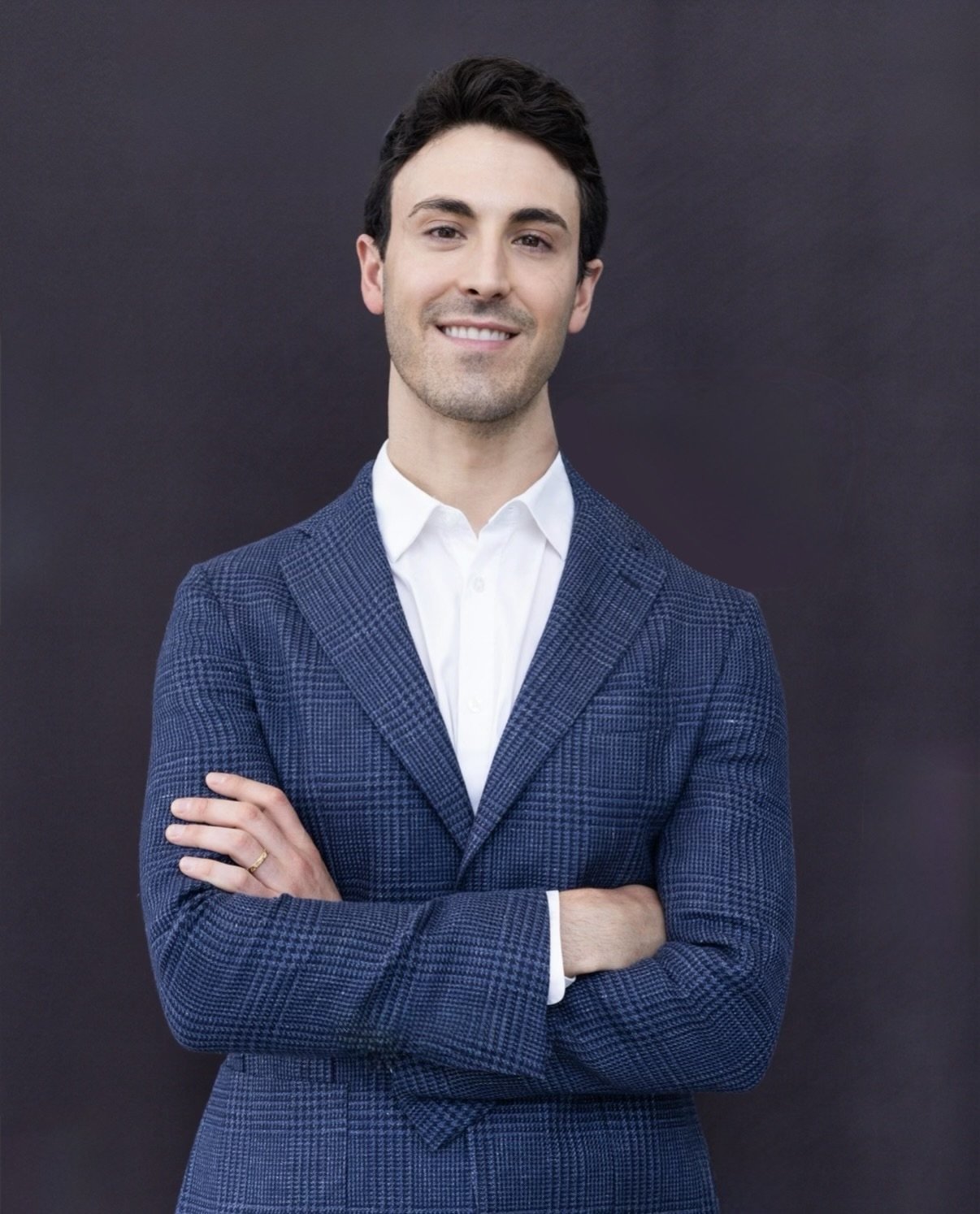 A young man in a blue checkered blazer and white shirt, smiling with arms crossed, standing against a dark background.