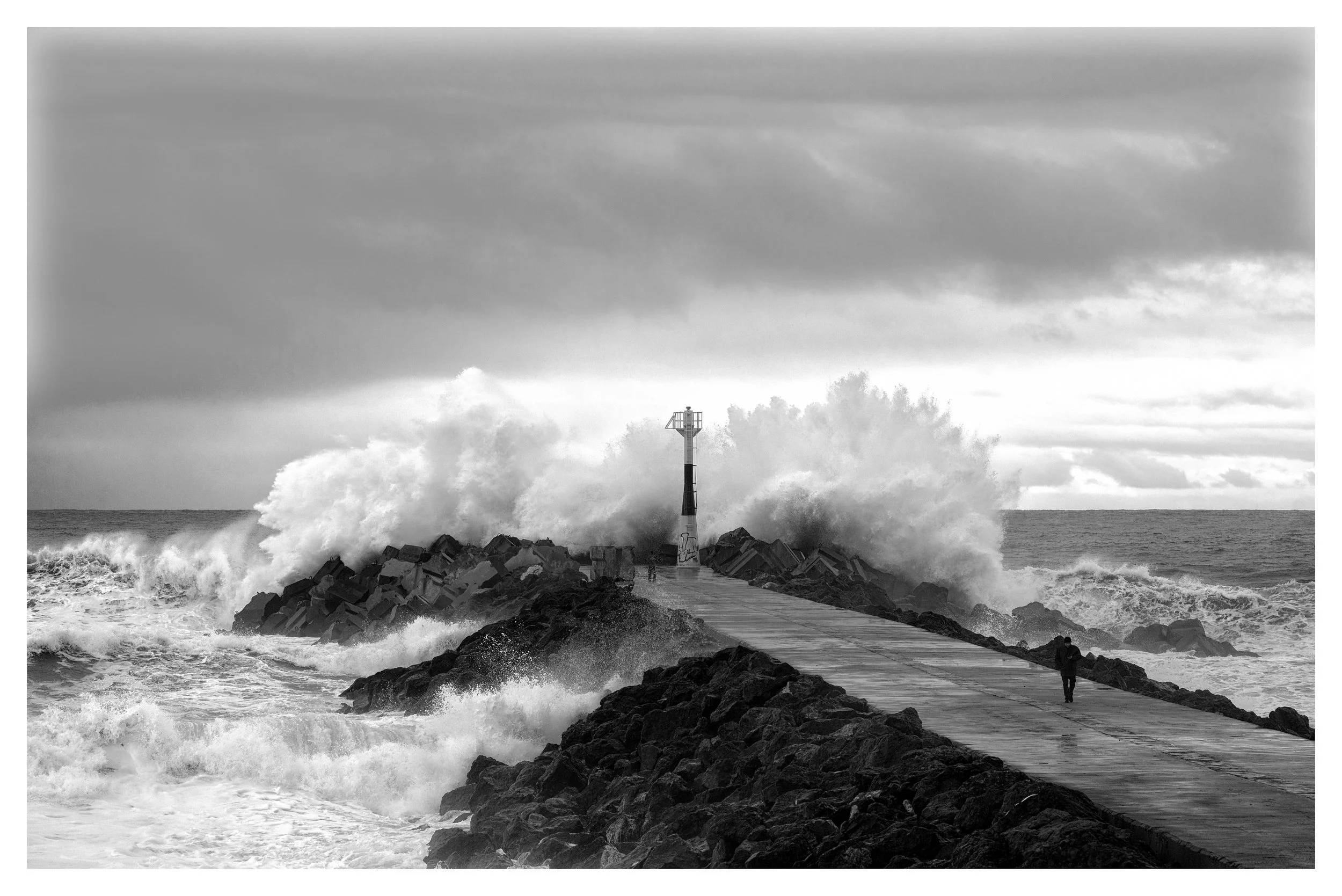 Une jetée en pierre s'étend dans la mer, avec des vagues s'écrasant contre elle. Un phare se trouve au bout de la jetée, tandis qu'une personne marche le long du quai par temps nuageux.