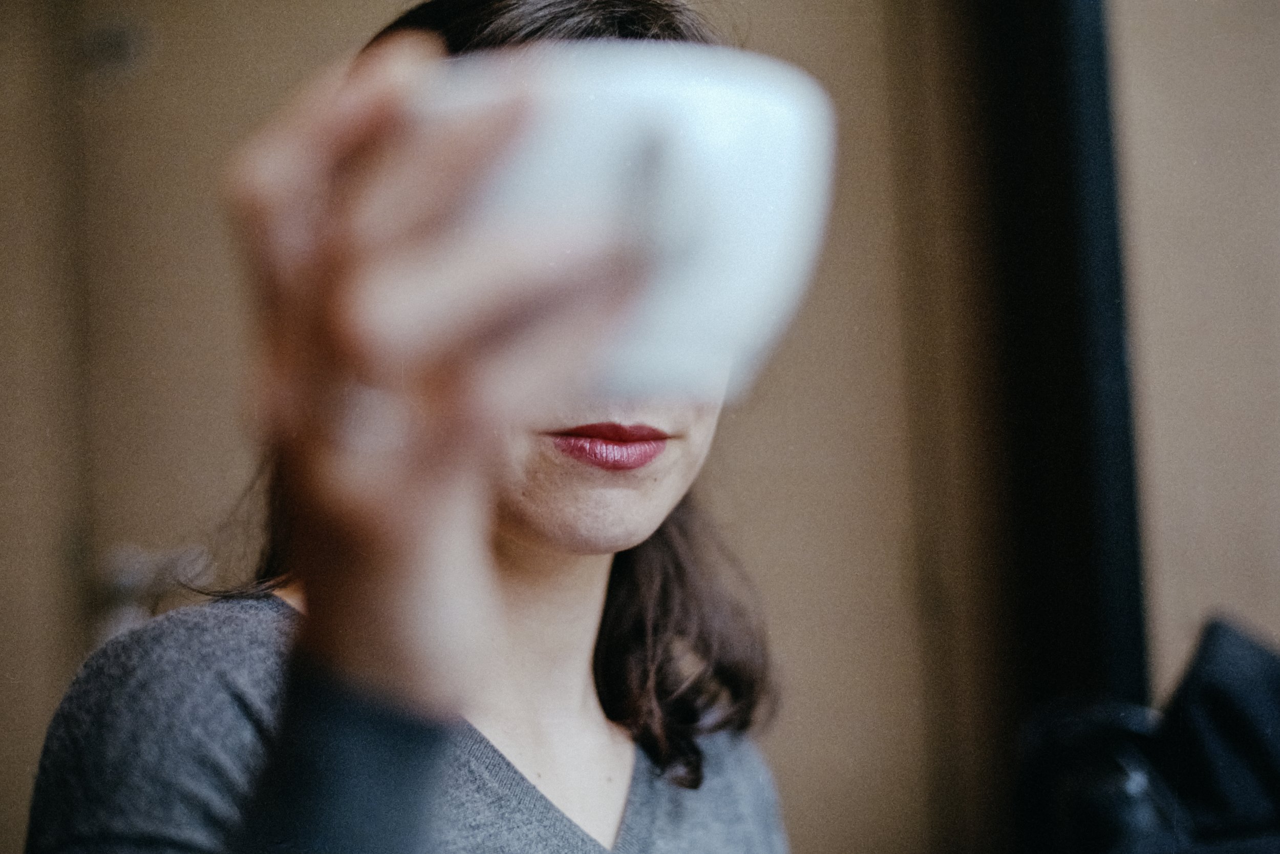 Une femme avec des cheveux bruns, portant un pull gris, se regarde dans un miroir en prenant une photo d'elle-même. Son visage est partiellement visible, avec un maquillage discret, dans un cadre intérieur.