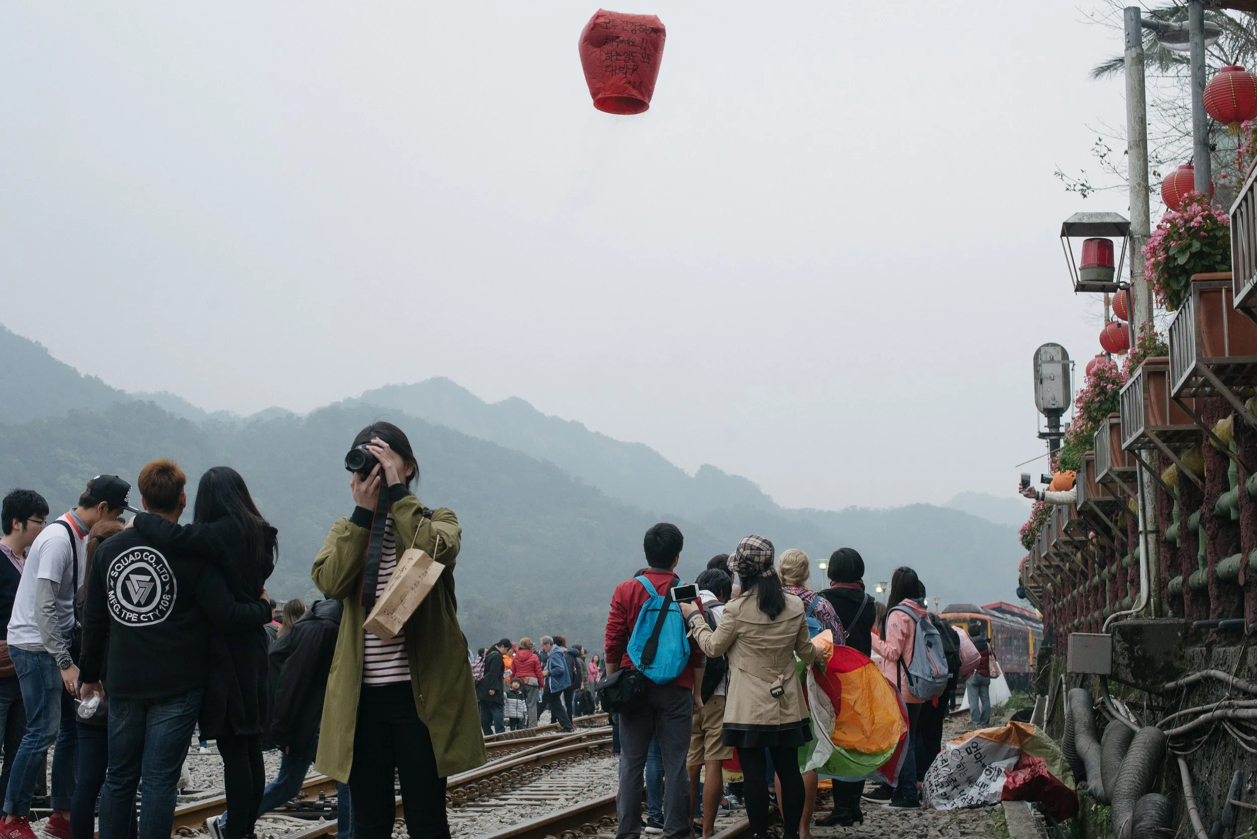 Une foule de personnes rassemblées près de lignes de chemin de fer, des montagnes en arrière-plan, un ciel nuageux, et des lanternes rouges suspendues, avec des bâtiments ornés de fleurs sur le côté droit.