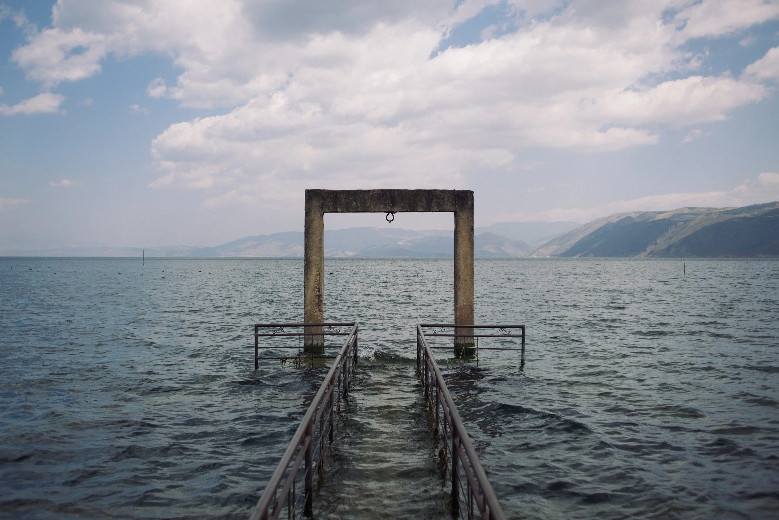 Une structure en béton avec une porte rectangulaire suspendue au-dessus de l'eau, probablement sur un lac ou une mer, avec des montagnes en arrière-plan et un ciel partiellement nuageux.