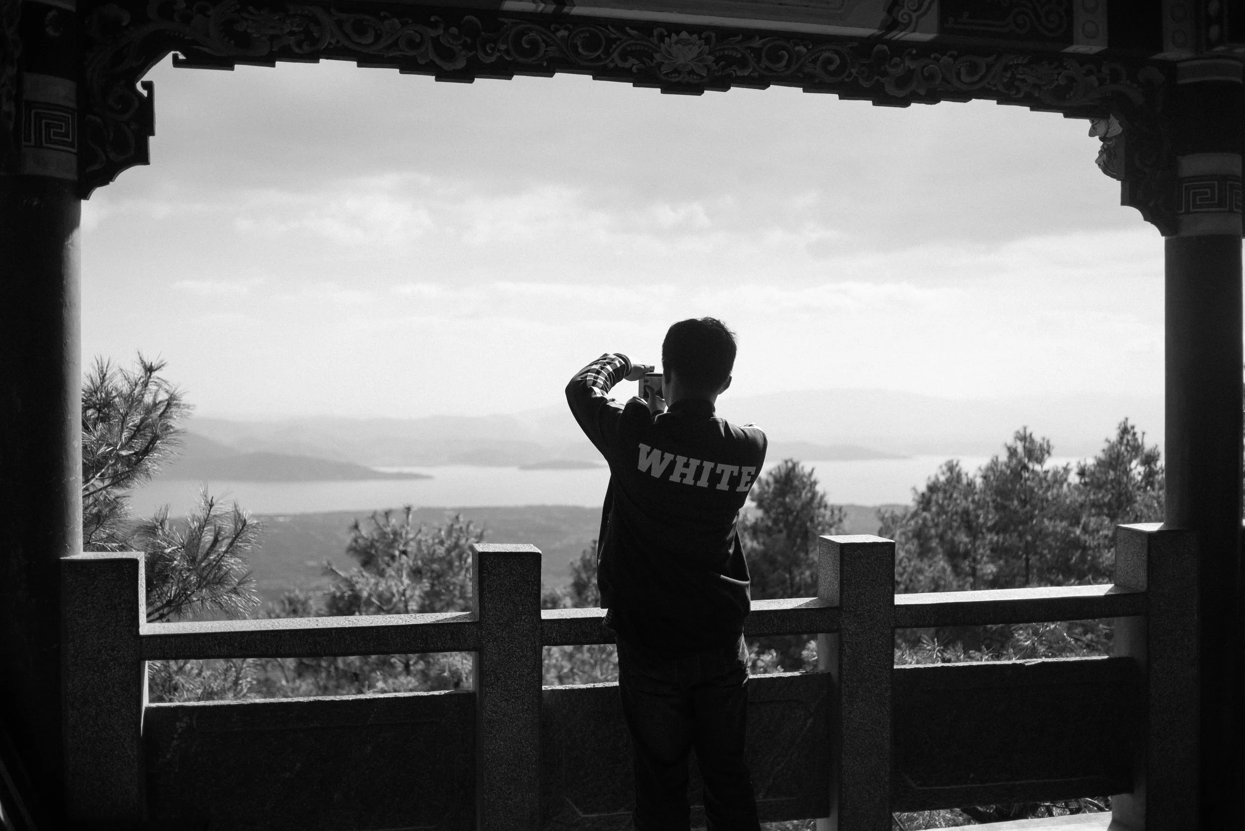Un homme en t-shirt noir avec le mot "WHITE" à l'arrière, prenant une photo avec un téléphone devant un paysage en montagne, vu à travers une structure en bois avec des colonnes et un toit décoré.