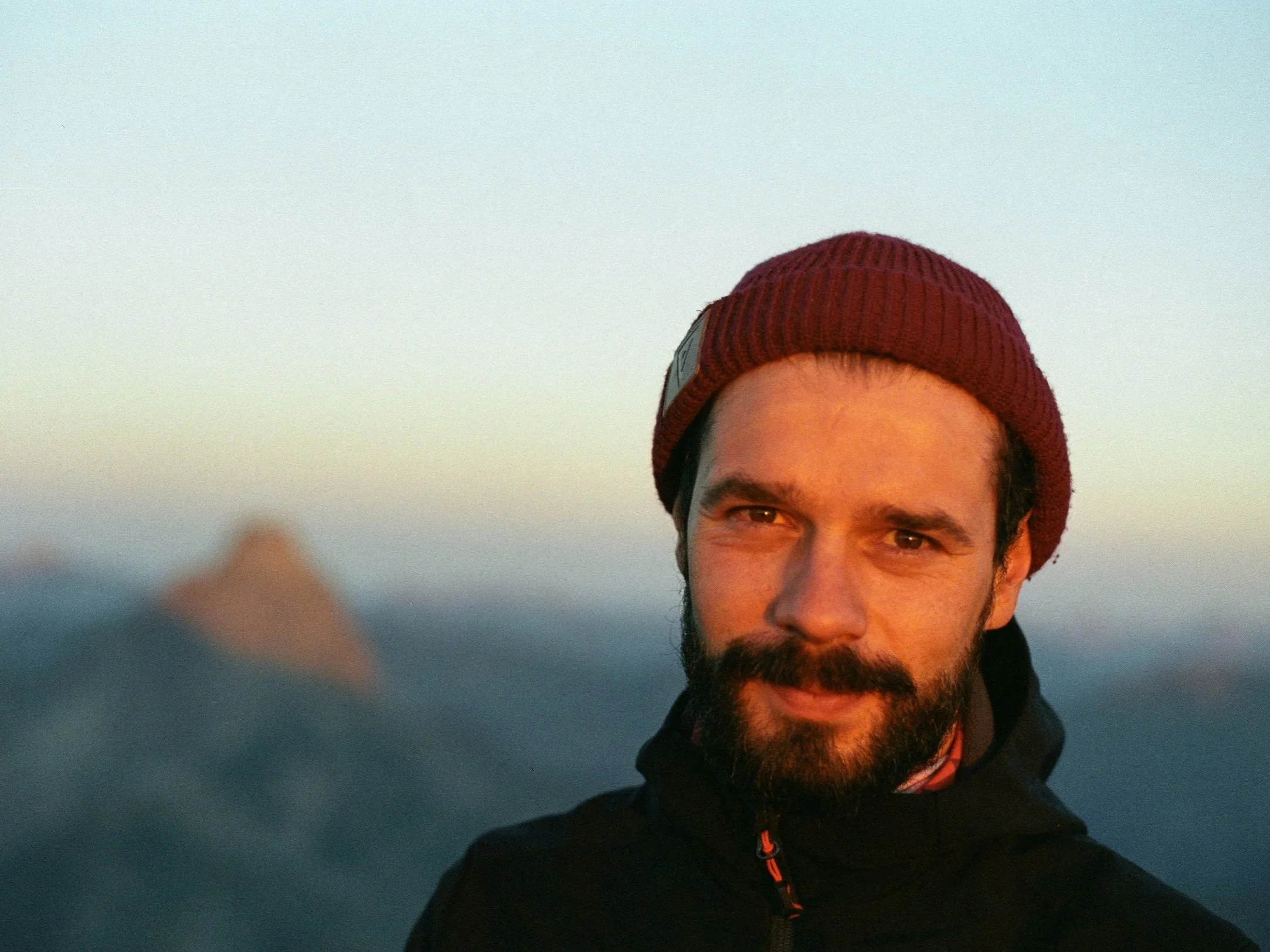 Portrait d'un homme portant un bonnet rouge, souriant légèrement, avec un fond de ciel clair au crépuscule.