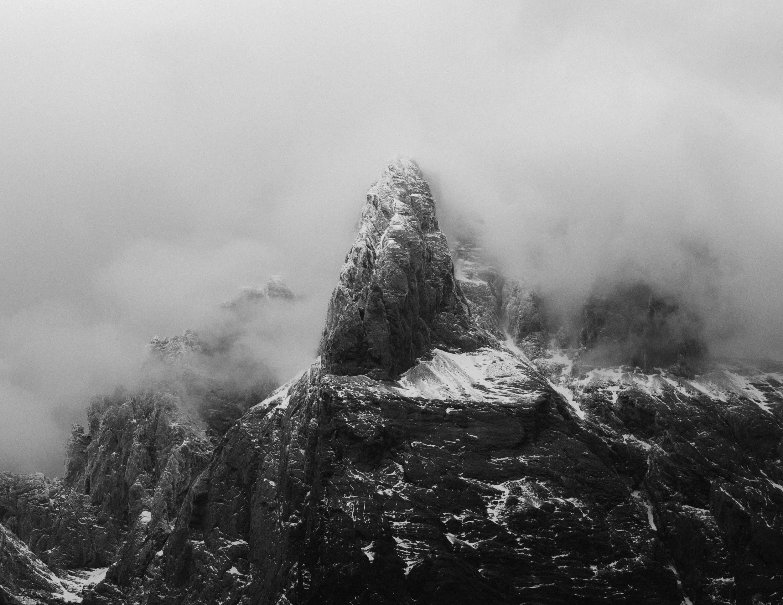 Une montagne enneigée enveloppée de nuages et de brouillard, avec des rochers imposants au premier plan en noir et blanc.