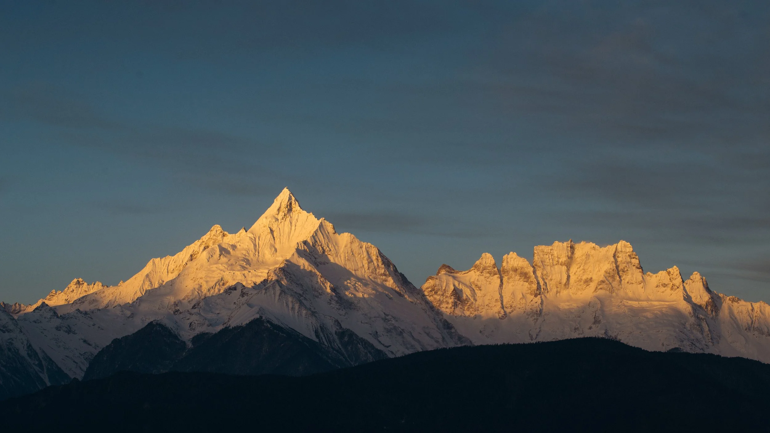 Montagnes enneigées au lever ou coucher du soleil avec un sommet principal pointu et des nuages au ciel.