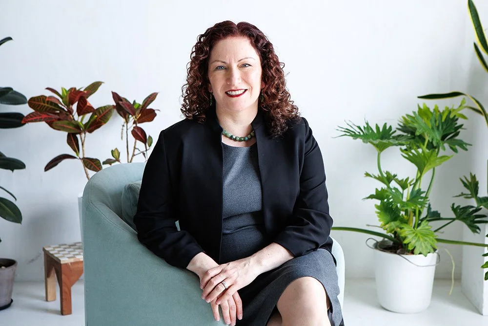 Photo of Anna Matas, lawyer and founder of Matas Legal. Sitting on a light blue chair surrounded by green plants.