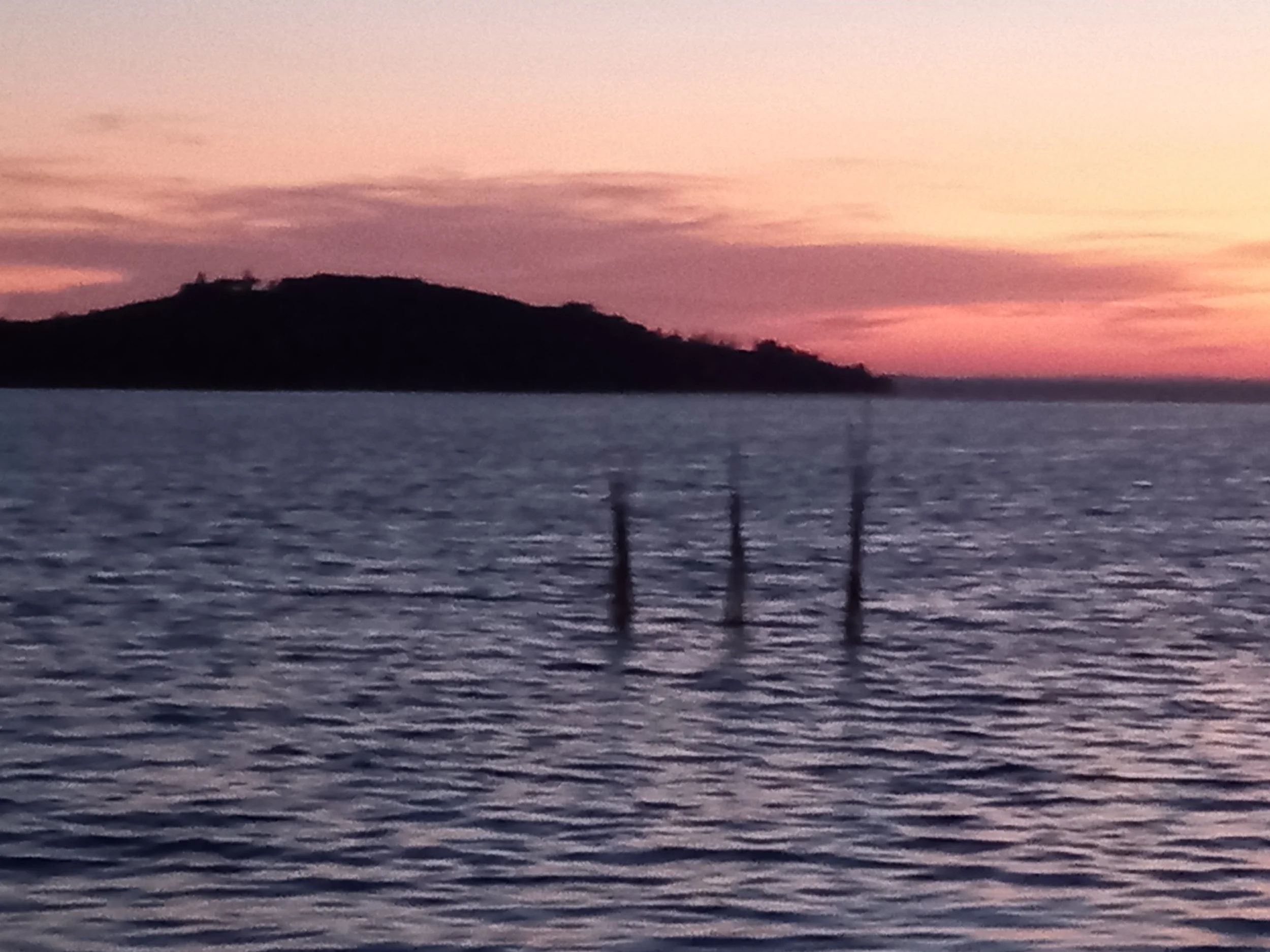 The image shows a scenic view of a body of water during sunset with colorful pink, purple, and orange sky. In the distance, there is a silhouette of a hill or small mountain with some trees or structures on top. The foreground features some dark vertical posts or poles along the water's edge.