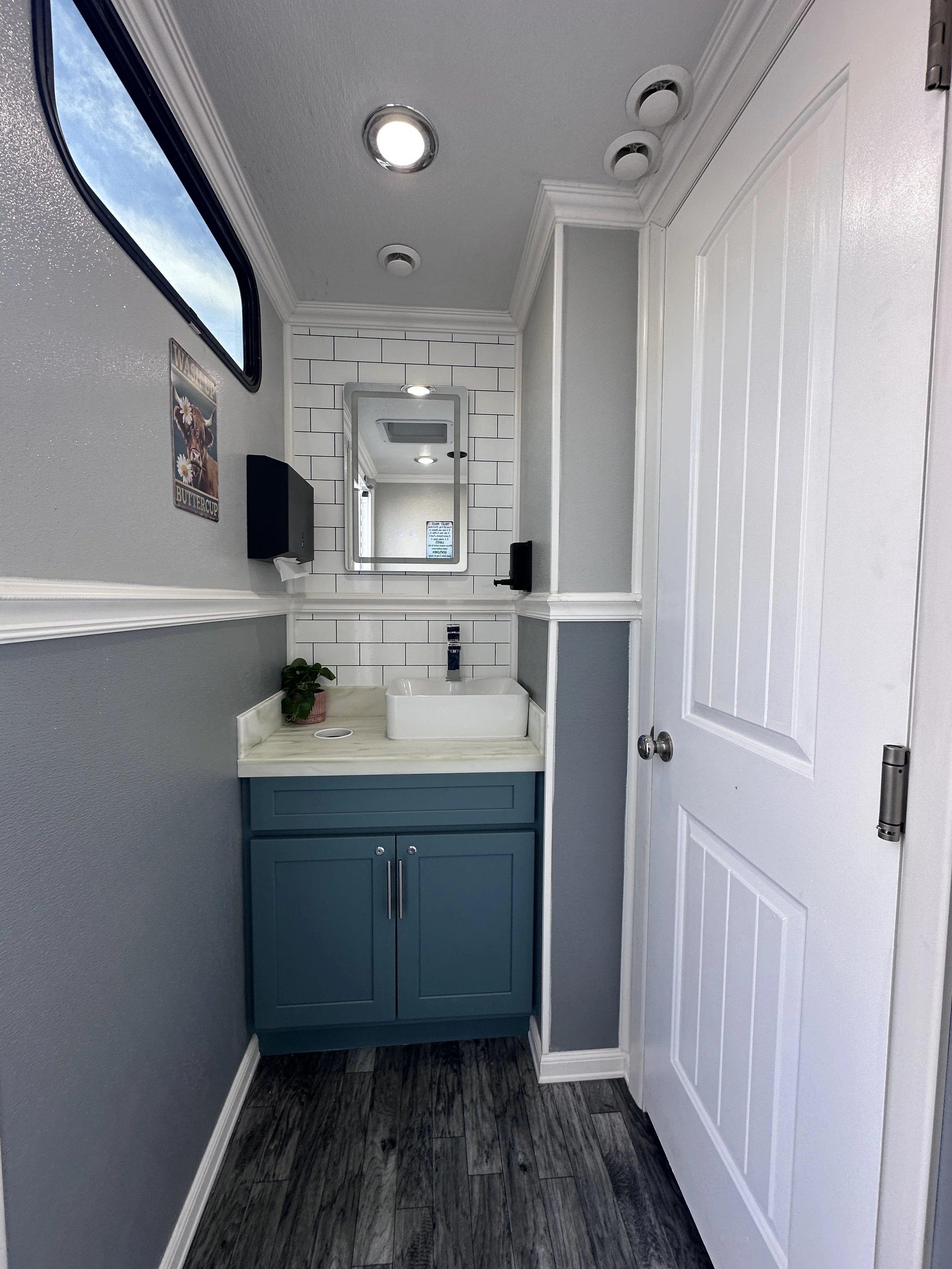 A small bathroom with gray walls, a white beadboard cabinet, a white countertop sink, a mirror, a soap dispenser, a plant, a window showing a blue sky, and ceiling lights.