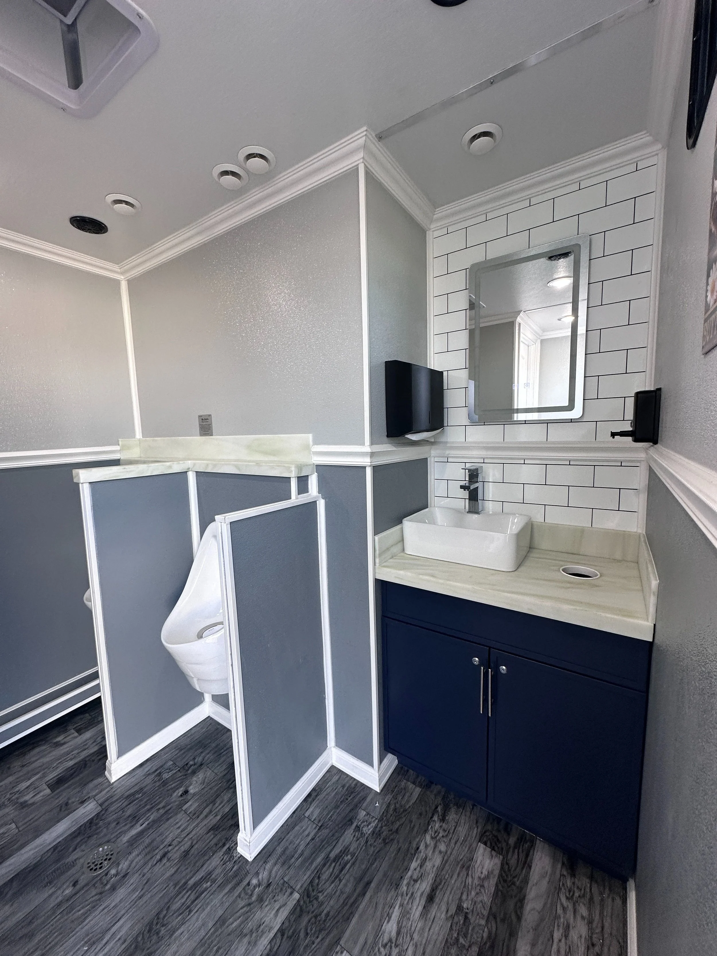 Public restroom with a sink, mirror, soap dispenser, and partial stalls with gray partitions, on dark wooden flooring.