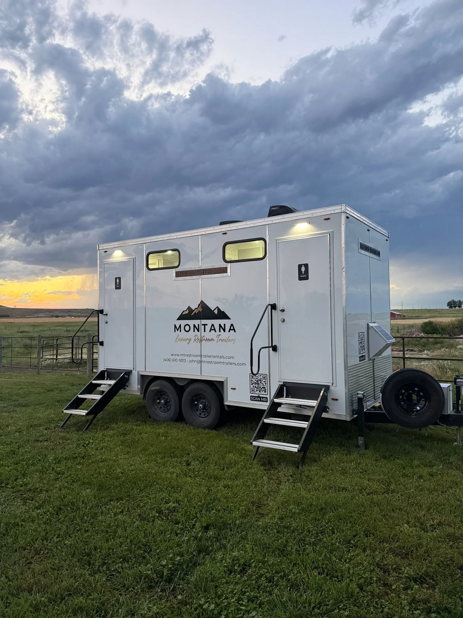 A white luxury restroom trailer with steps leading up to the door, located outdoors on a grassy area with a cloudy sky overhead. The trailer is branded with a Montana mountain logo and website information.