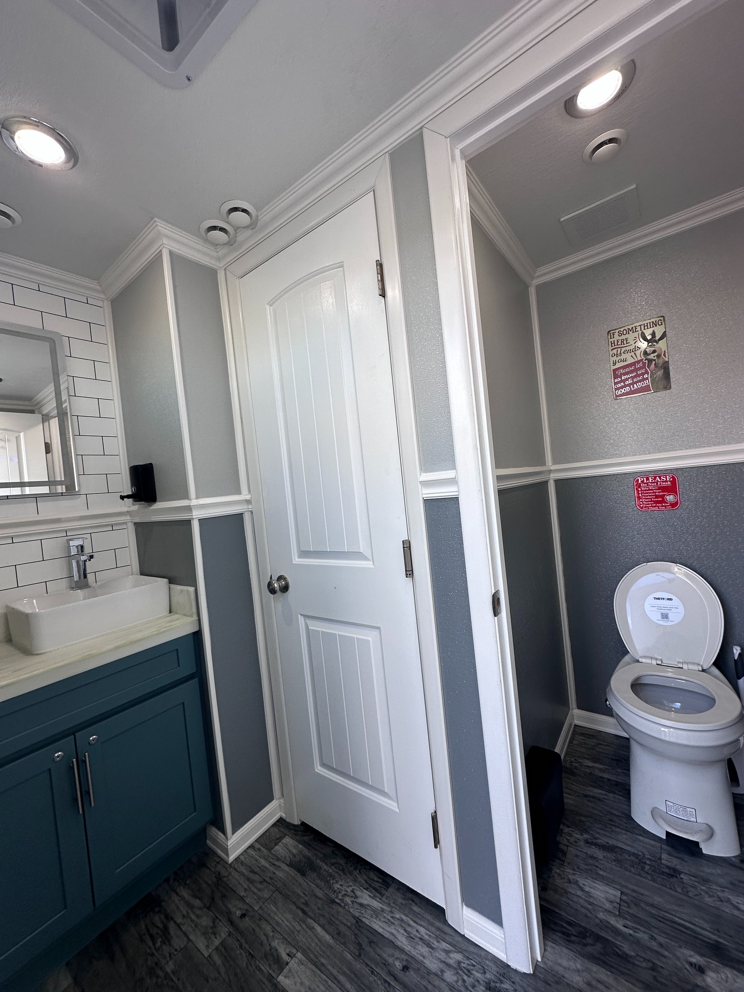Bathroom with white door, gray walls, blue vanity with sink, white subway tiles, ceiling lights, a mirror, and a white toilet. There are signs on the wall, one says 'If something here offends you please let us know so you can all use a good laugh.'