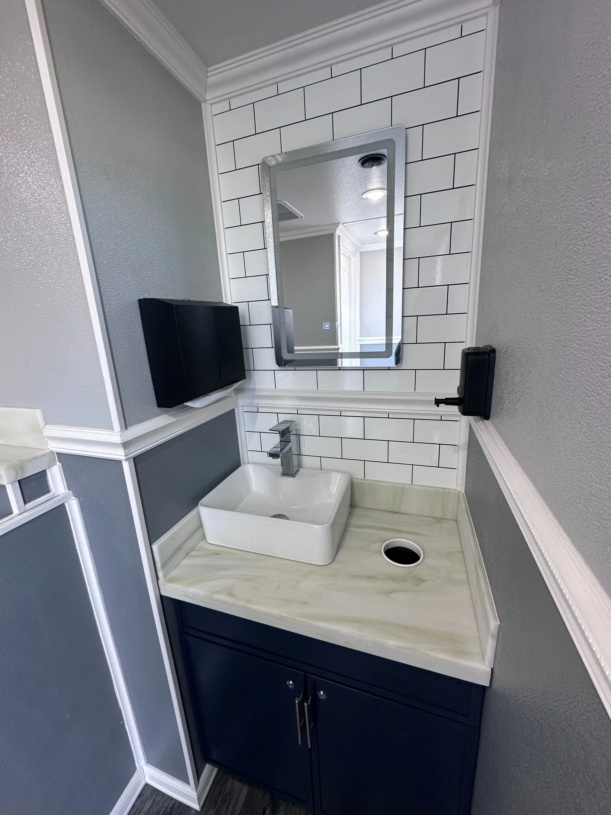 A bathroom vanity with a white vessel sink, a faucet, a small mirror, black soap dispenser, dark blue cabinetry, and a white tile backsplash with black grout.