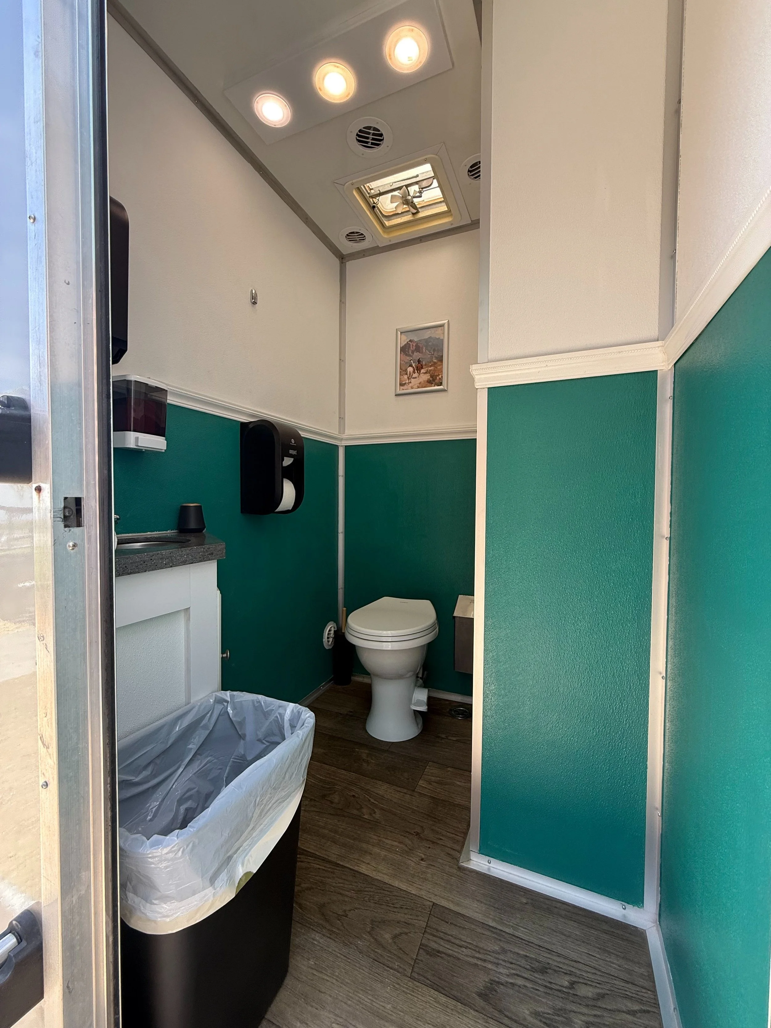 Compact restroom with a toilet, a trash can, privacy divider, a small sink, and a soap dispenser on the wall.
