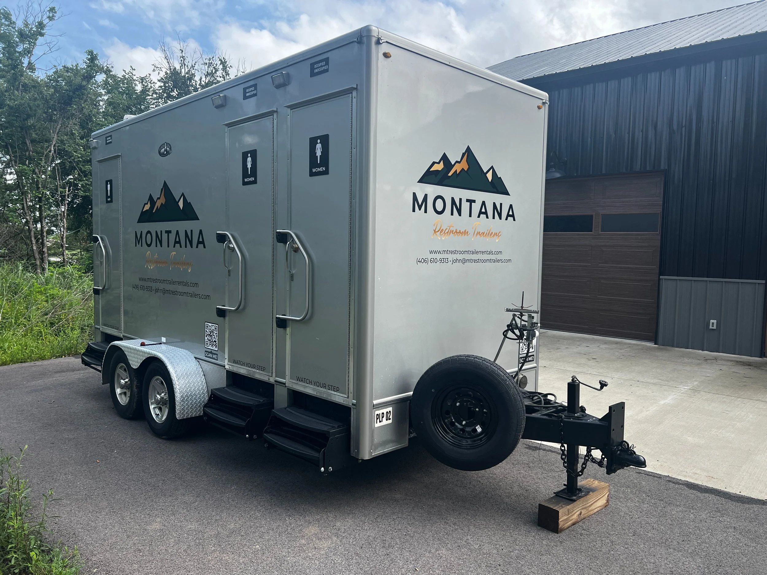 A white trailer with mountain and Montana logo, placed on a paved area next to a black building with a metal roof, trees in the background, and a partly cloudy sky.