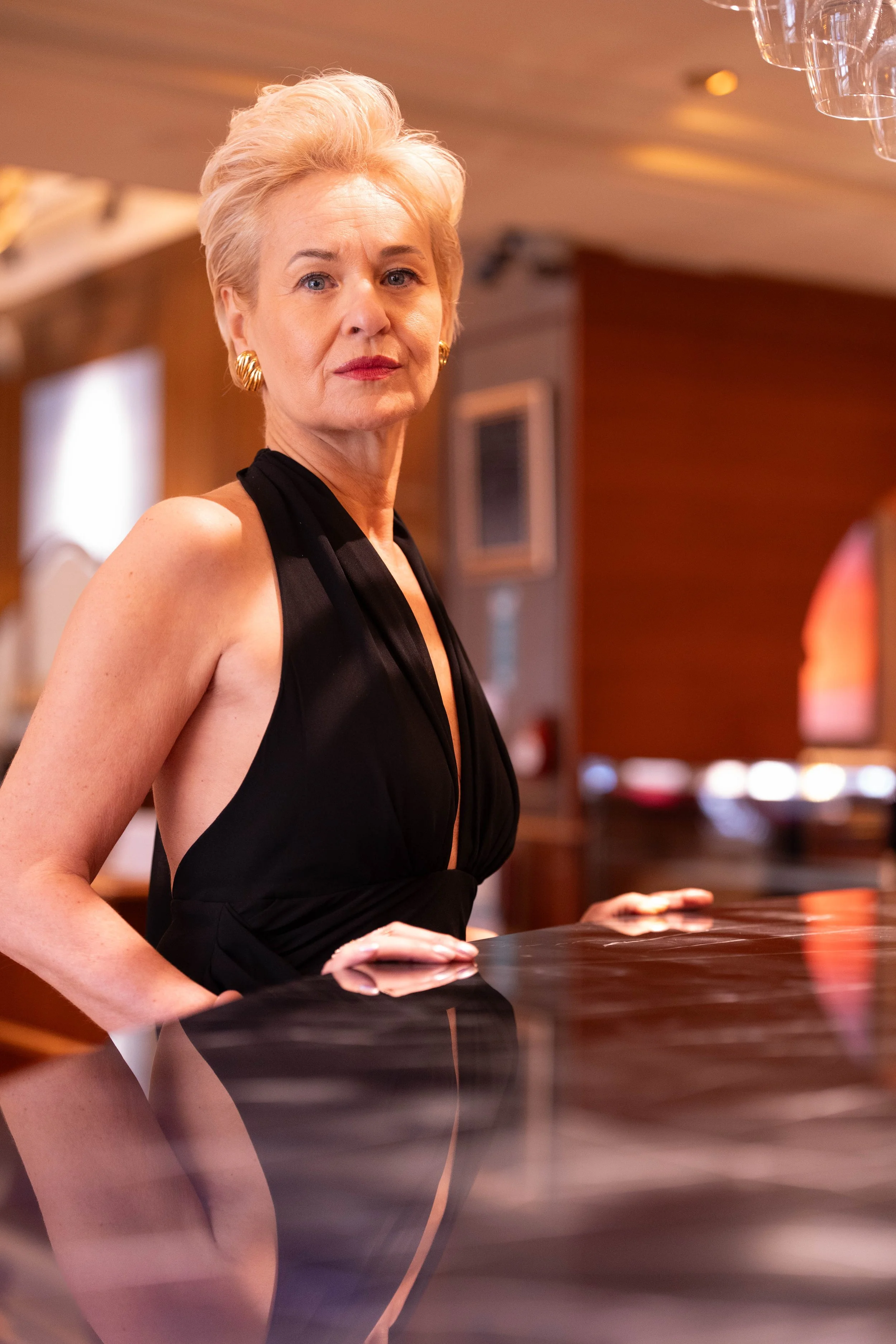Une femme élégante avec des cheveux blonds courts, portant une robe noire sophistiquée, posant dans un lieu intérieur avec un décor chaud.