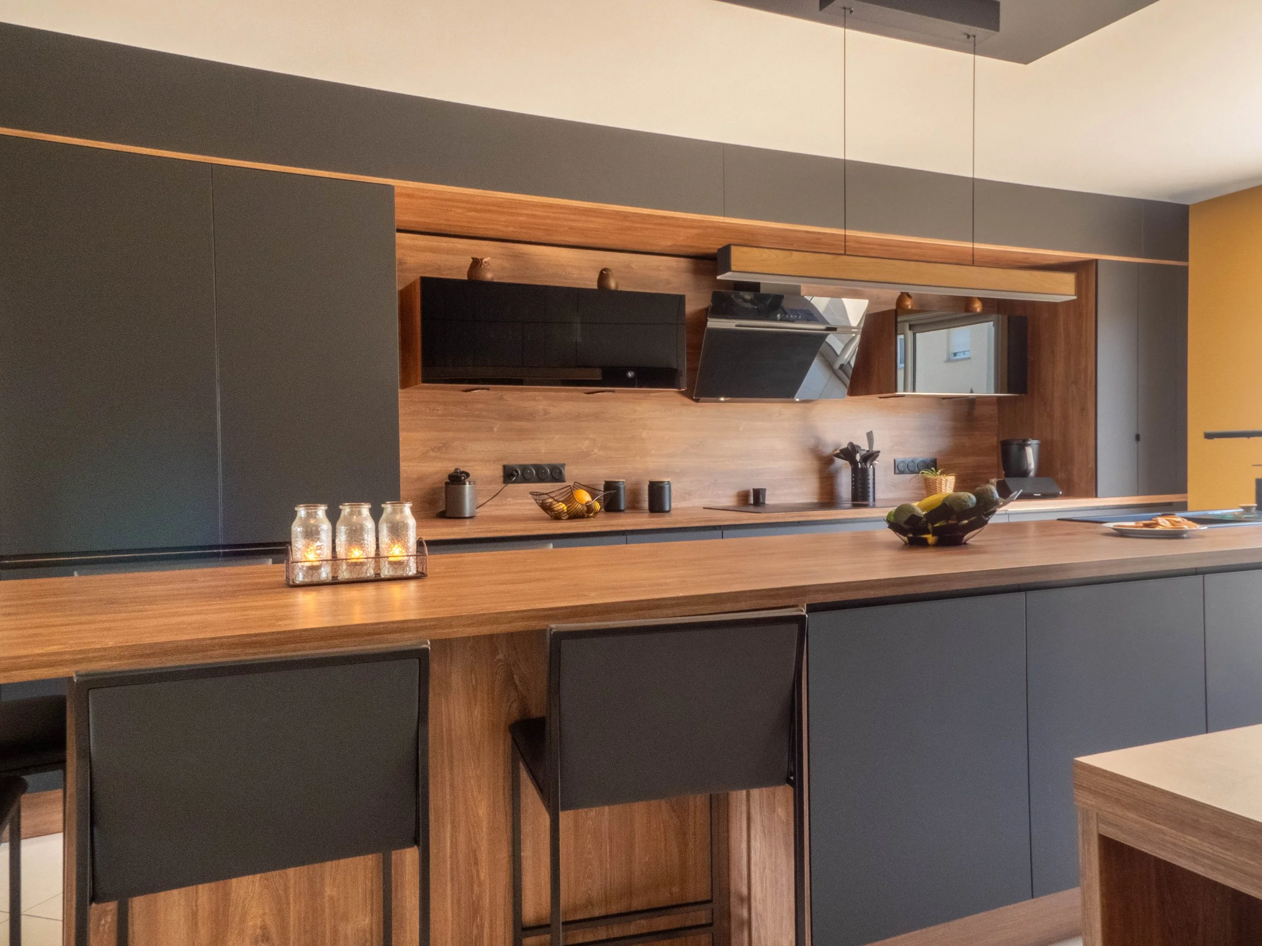Cuisine moderne avec une finition en bois, étagères et appareils électroménagers noirs, un plan de travail en bois avec des lampes en verre, un bol de fruits et des ustensiles.