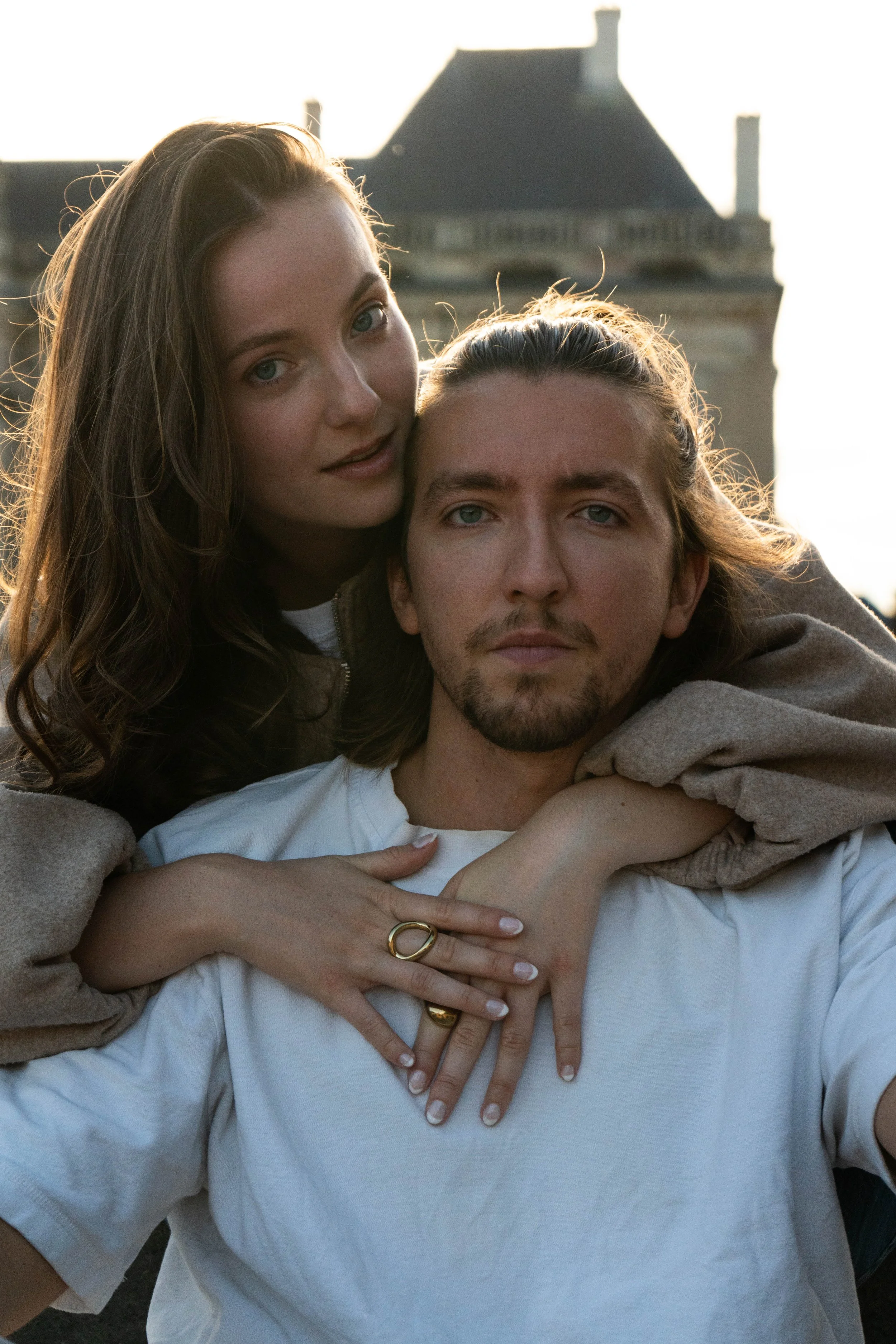 Un couple posant à l'extérieur au coucher du soleil, la femme se tenant derrière l'homme, tous deux regardant vers la caméra, avec une maison en arrière-plan.