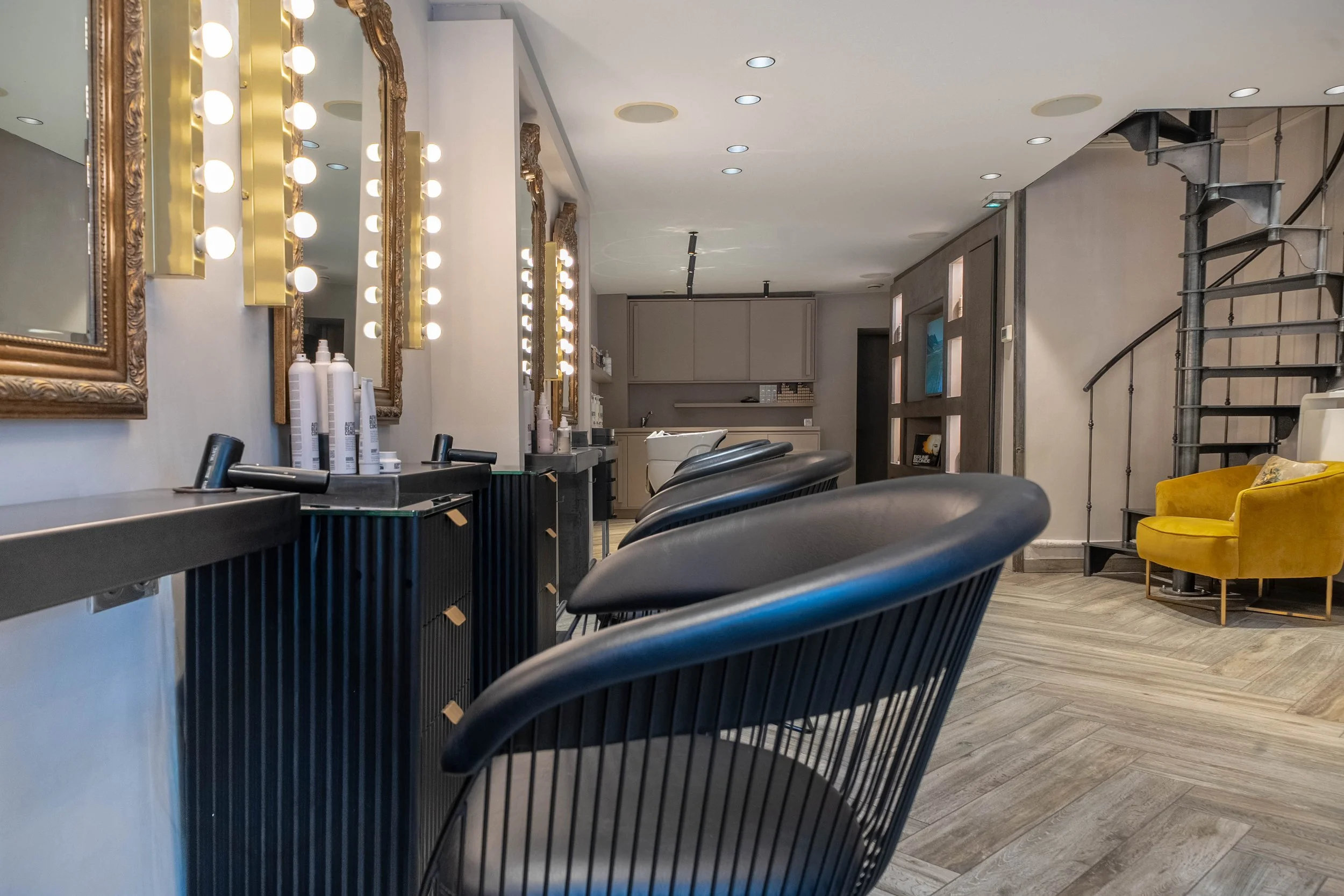 Salon de coiffure avec des miroirs encadrés de doré, des luminaires à ampoules, des fauteuils de coiffure noirs, une étagère avec des produits capillaires, un mur avec une télévision, un escalier en métal, et un fauteuil jaune.