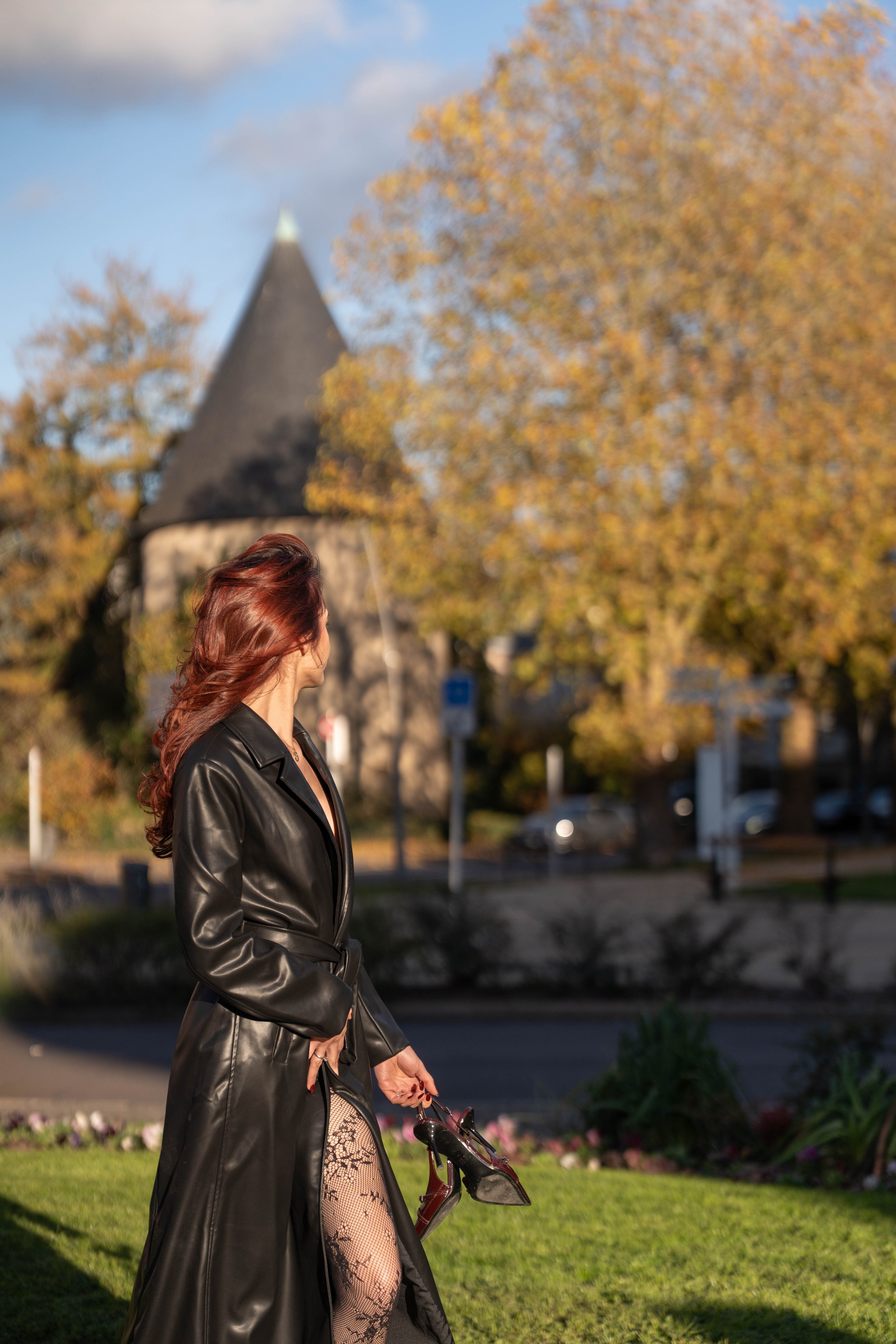 Une femme aux cheveux roux portant un manteau en cuir noir marche dans un parc avec un arbre aux feuilles automnales en arrière-plan, un château ou une vieille tour en pierre est visible en arrière-plan.