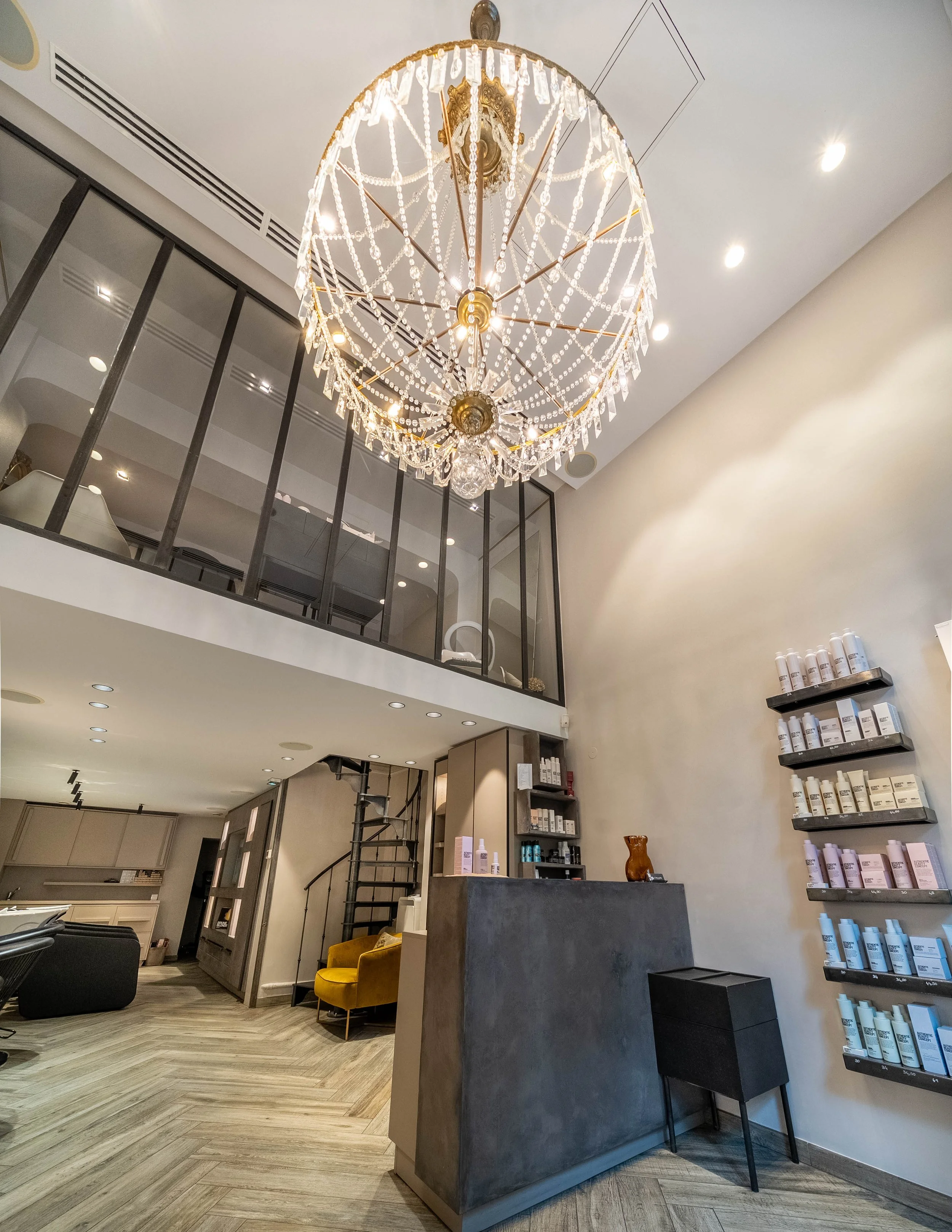Intérieur d'un salon ou d'un spa avec un chandelier en cristal suspendu au plafond, un comptoir en pierre grise, des étagères avec des produits de soins, une chaise noire, un fauteuil jaune, et une disposition moderne avec un sol en bois.