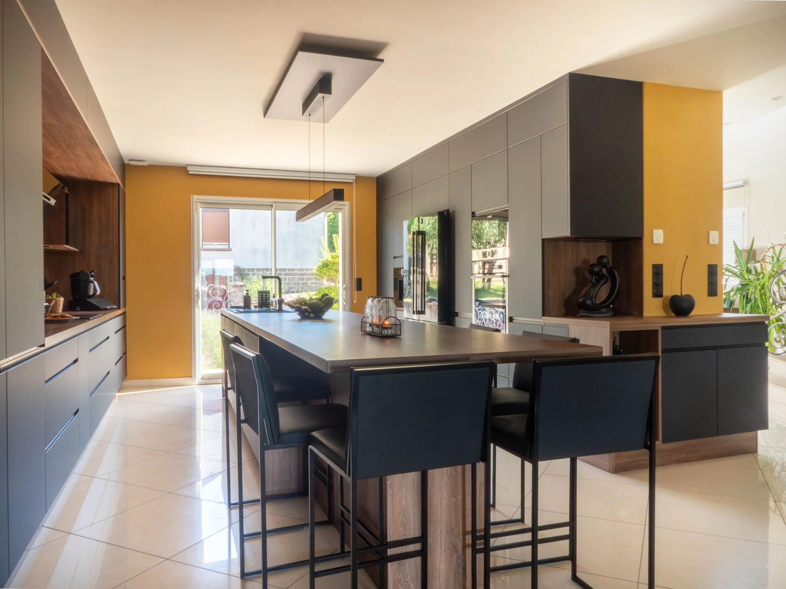 Cuisine moderne avec un îlot au centre, mobilier en bois et placards gris clair, grandes fenêtres laissant entrer la lumière naturelle, décoration épurée avec quelques objets décoratifs