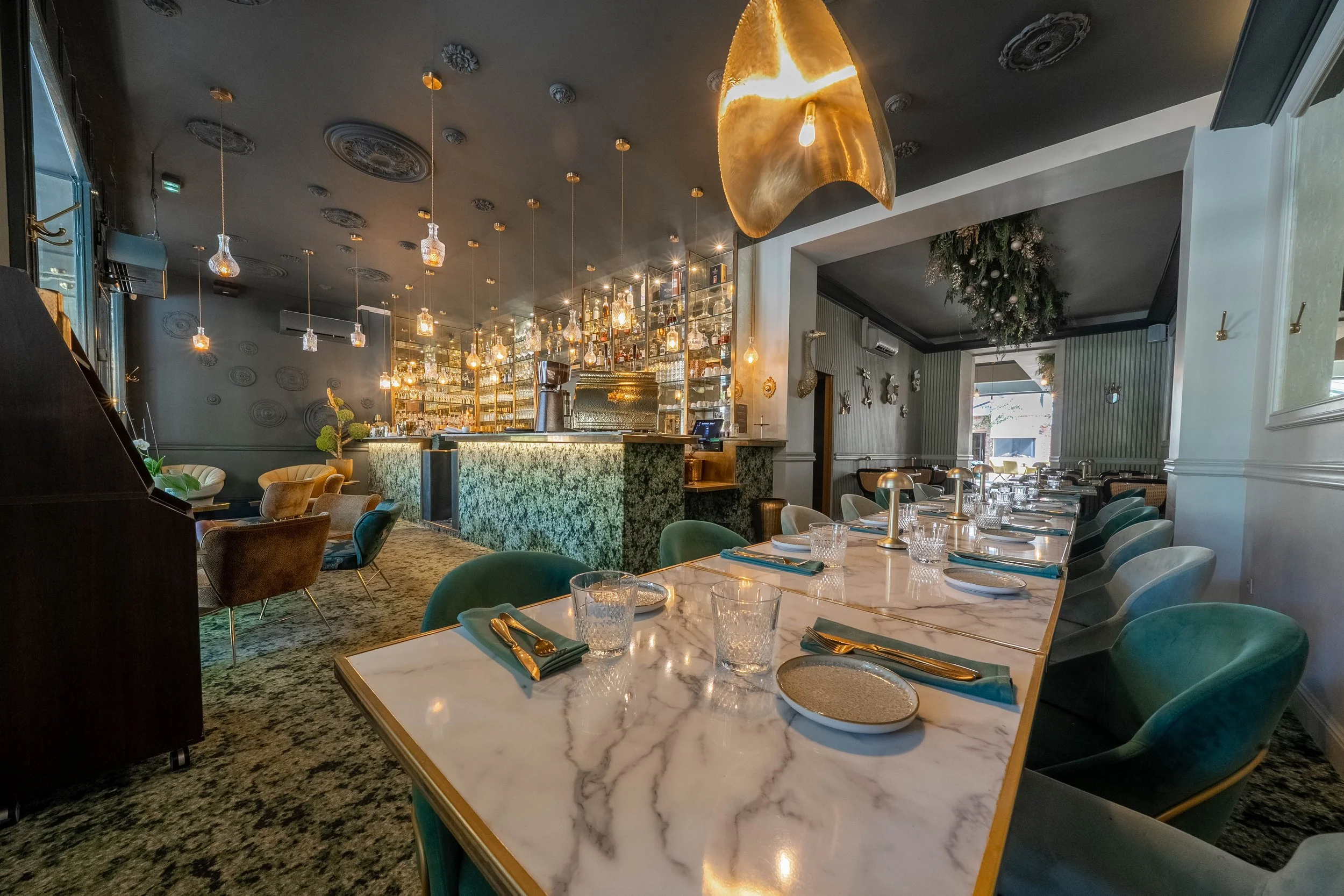 Intérieur d'un restaurant élégant avec un bar en arrière-plan, des tables avec vaisselle et couverts, des sièges en velours vert et beige, une lumière suspendue en forme de feuille dorée, et une décoration de Noël avec des branches de sapin suspendue