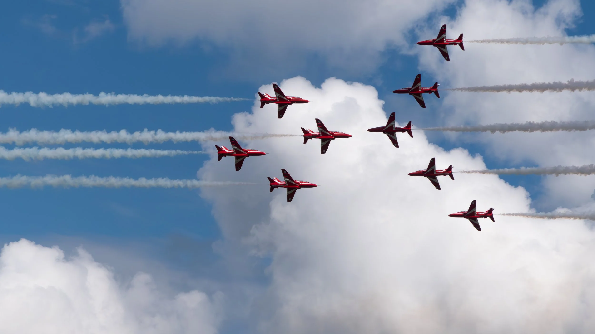 Seven avions rouges effectuant un spectacle aérien en formation dans le ciel avec des traînées de fumée blanche.