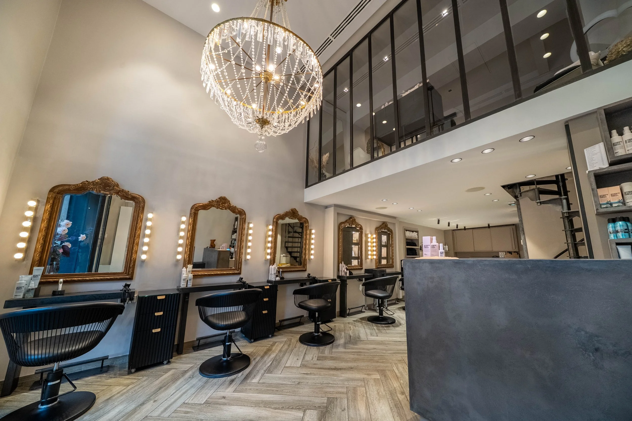 Salon de coiffure moderne avec plusieurs miroirs encadrés de lumière, fauteuils noirs, grande lampe en cristaux au plafond, et étagères avec des produits capillaires.