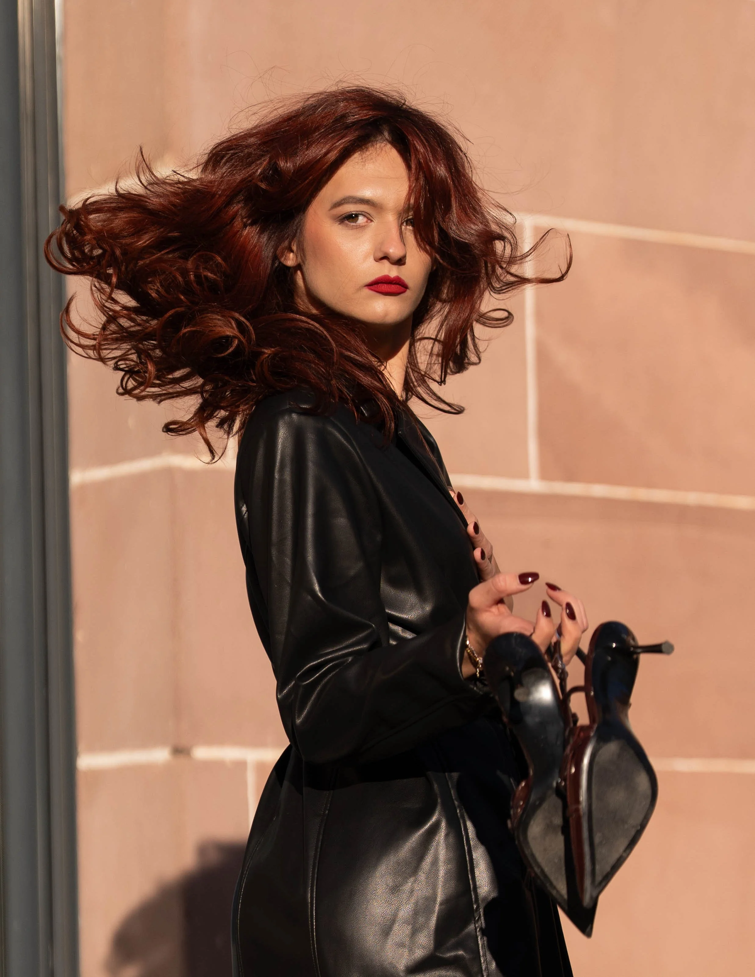 Femme aux cheveux roux bouclés, portant un maquillage rouge à lèvres, vêtue en cuir noir, se tenant contre un mur en pierre beige, regardant légèrement vers la caméra.