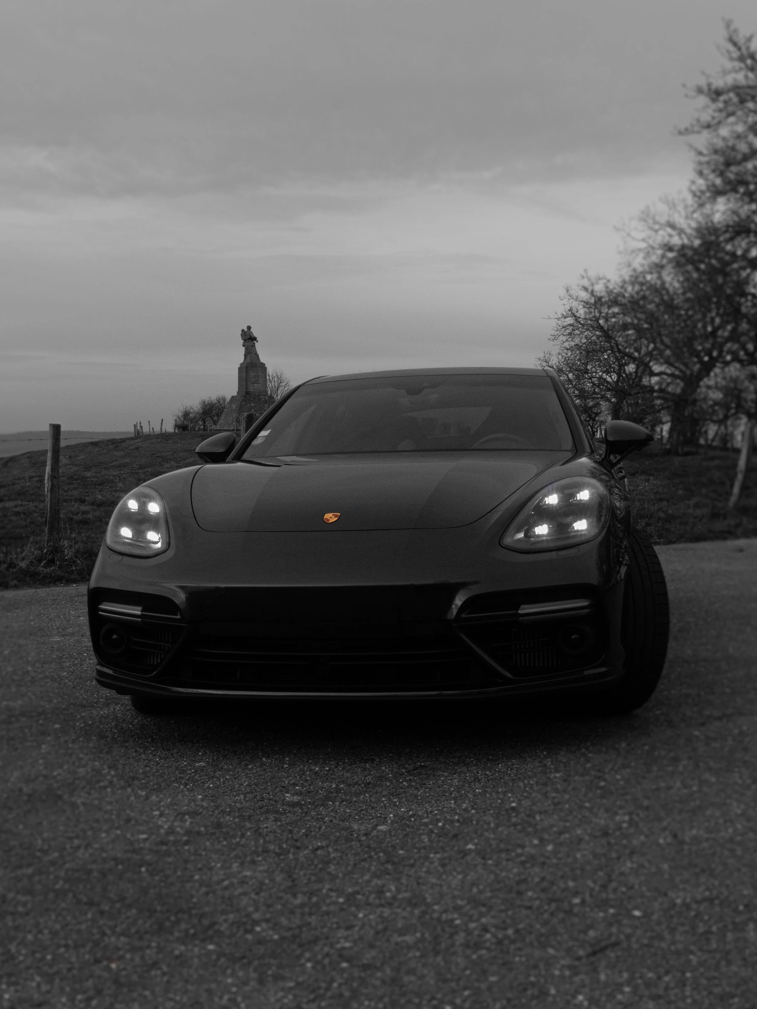Voiture Porsche noire garée sur une route avec un ciel nuageux, un monument en arrière-plan, et des arbres dénudés indiquant une saison hivernale ou automnale.