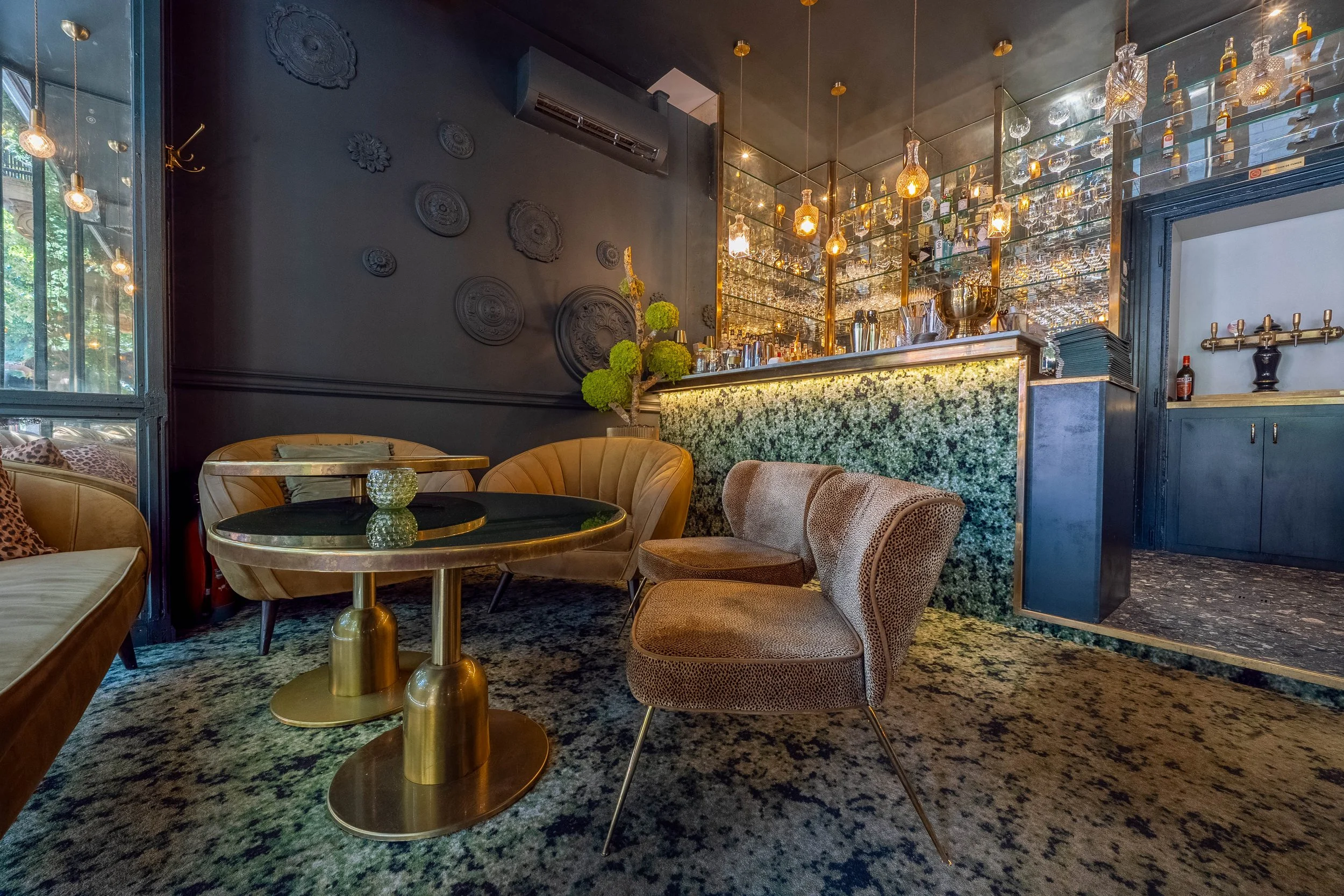 Interior d'un bar avec un comptoir en marbre vert, des lampes suspendues en verre et une décoration murale noire avec des motifs circulaires. Il y a des tables et des chaises, ainsi que des étagères avec des bouteilles d'alcool.