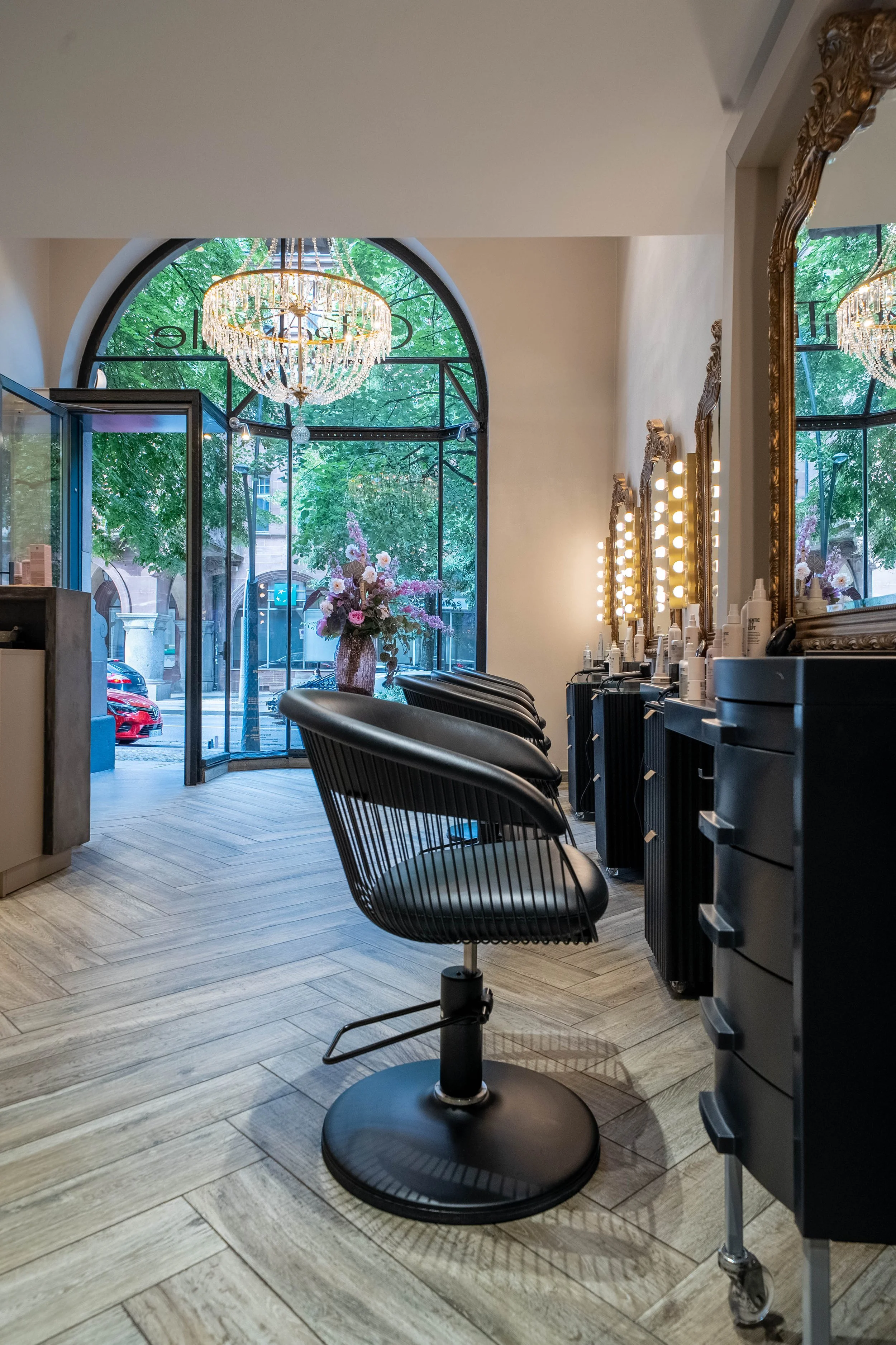 Intérieur d'un salon de coiffure avec une grande fenêtre, un lustre en cristal, des miroirs encadrés en or, des chaises de coiffure en bois noir, et des produits de soin alignés. Vue extérieure sur la rue et les arbres.