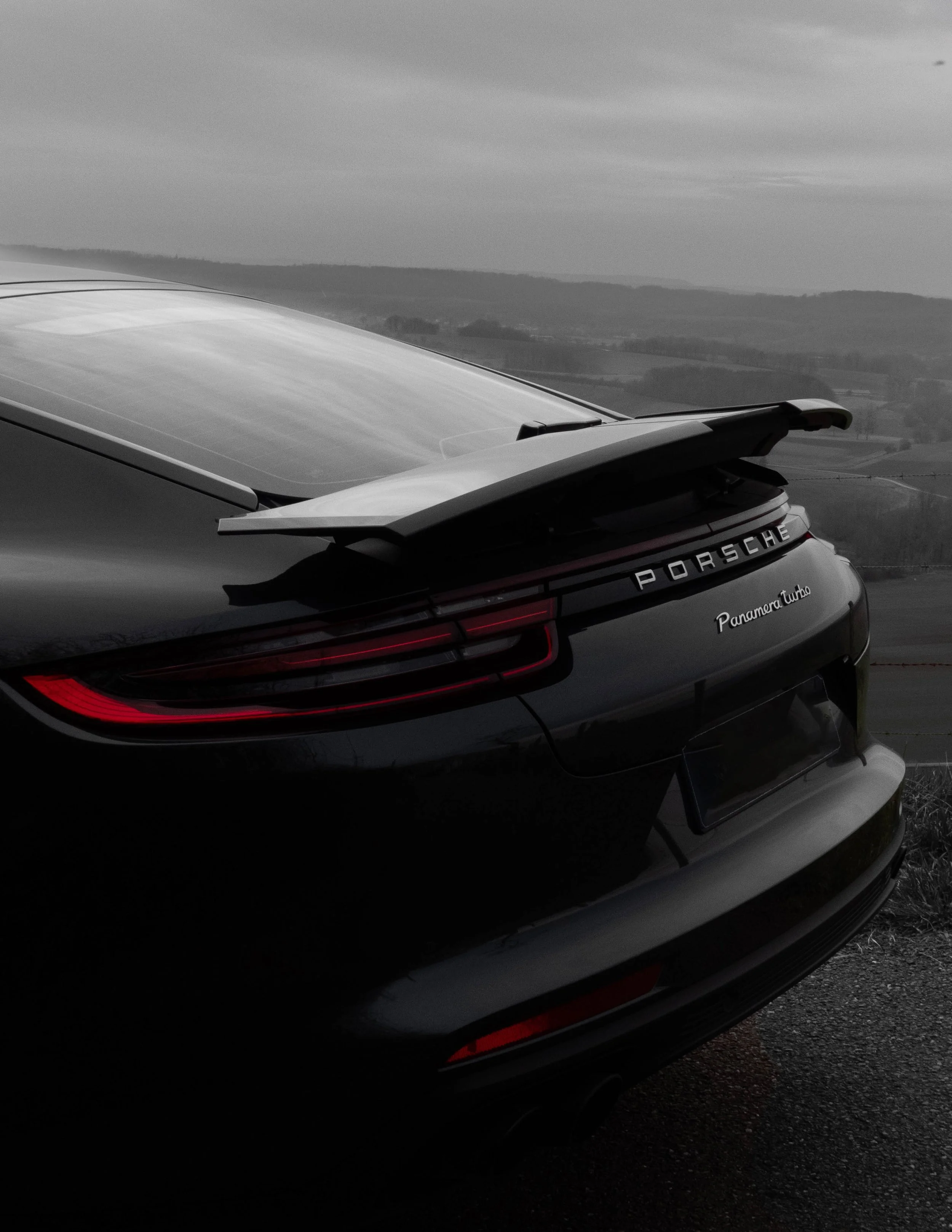 Vue partielle d'une voiture Porsche avec un aileron arrière, en cours d'observation dans un paysage rural avec un ciel nuageux.