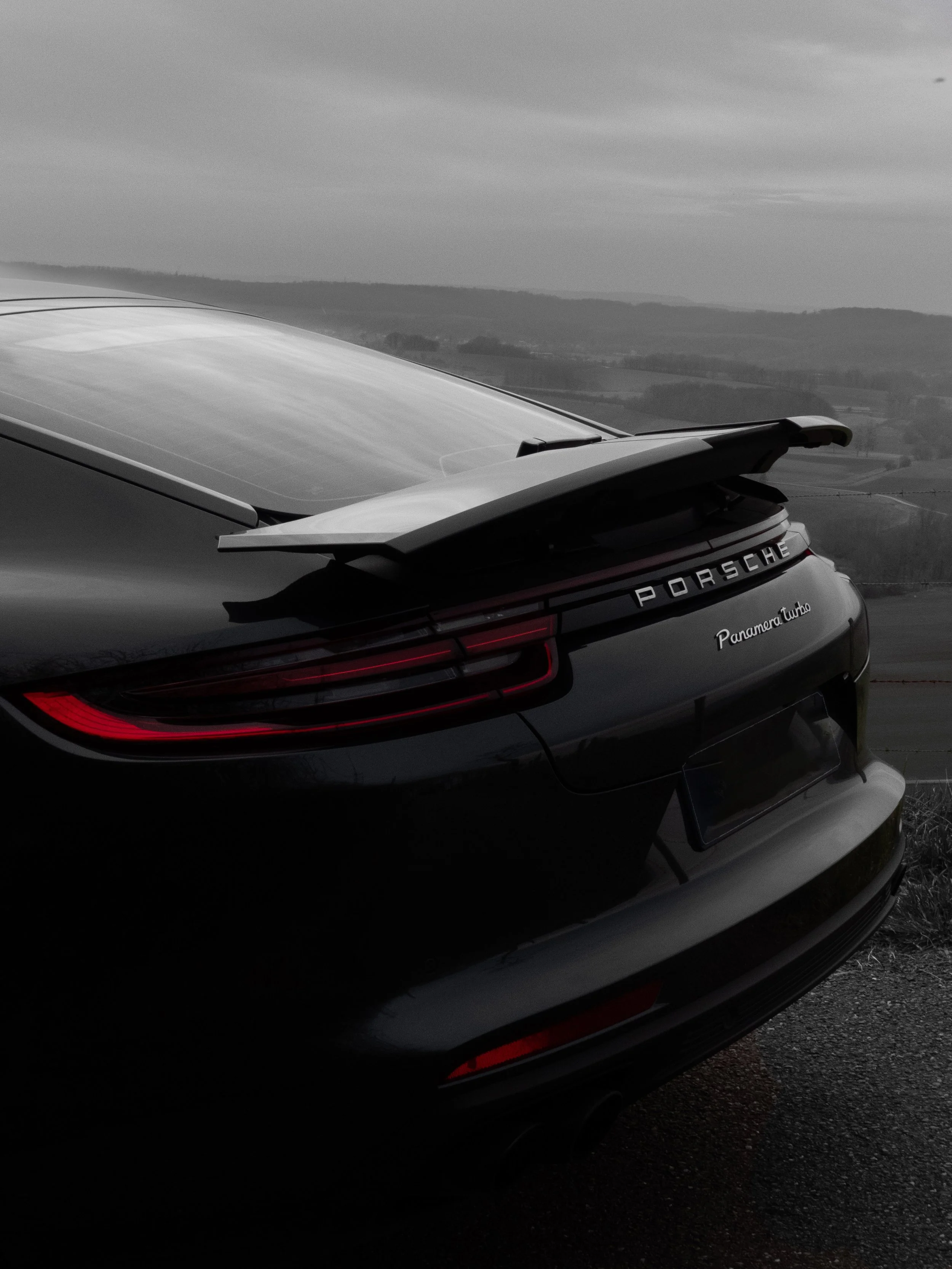 Vue arrière d'une voiture Porsche noire, modèle Panamera Turbo, avec un aileron arrière, stationnée sur une route rurale avec un paysage de collines et un ciel nuageux en arrière-plan.