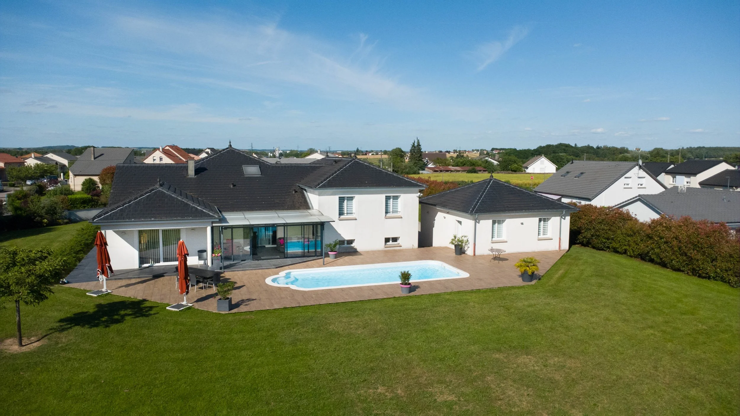Une maison moderne blanche avec une piscine extérieure entourée d'une terrasse en pierre, un jardin bien entretenu, des parasols rouges, et un paysage résidentiel avec d'autres maisons et de la verdure en arrière-plan.