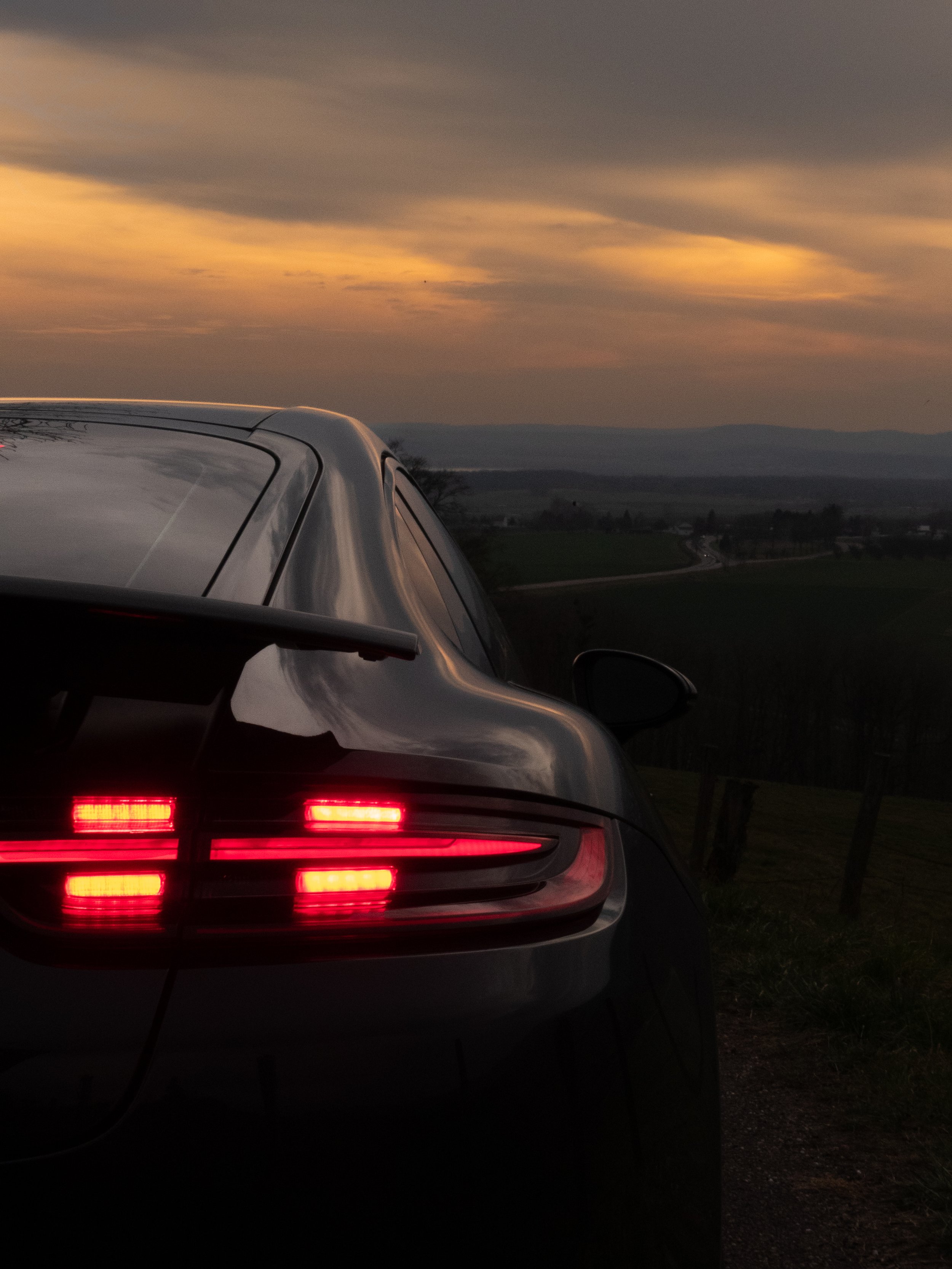Une voiture noire vue de l'arrière avec ses feux allumés, située dans un paysage rural au crépuscule, avec un ciel nuageux aux teintes orangées et un horizon vallonné.