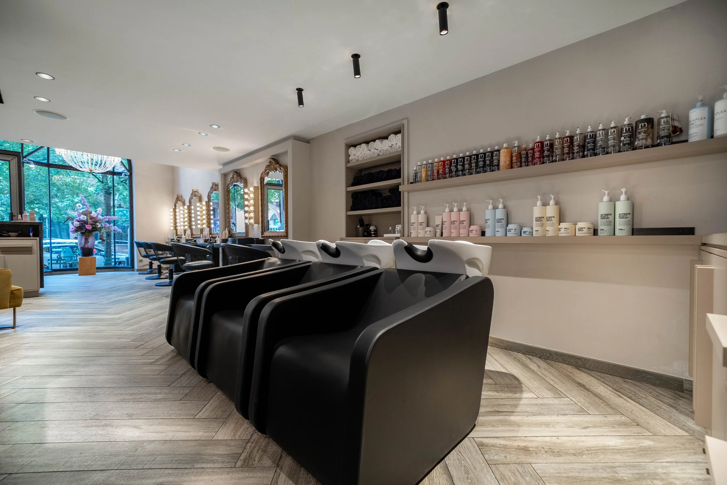 Une salle de salon de coiffure avec des lavabos noirs, des miroirs ornés de lumières, des étagères avec des serviettes et des produits capillaires, et une grande fenêtre laissant entrer la lumière naturelle.